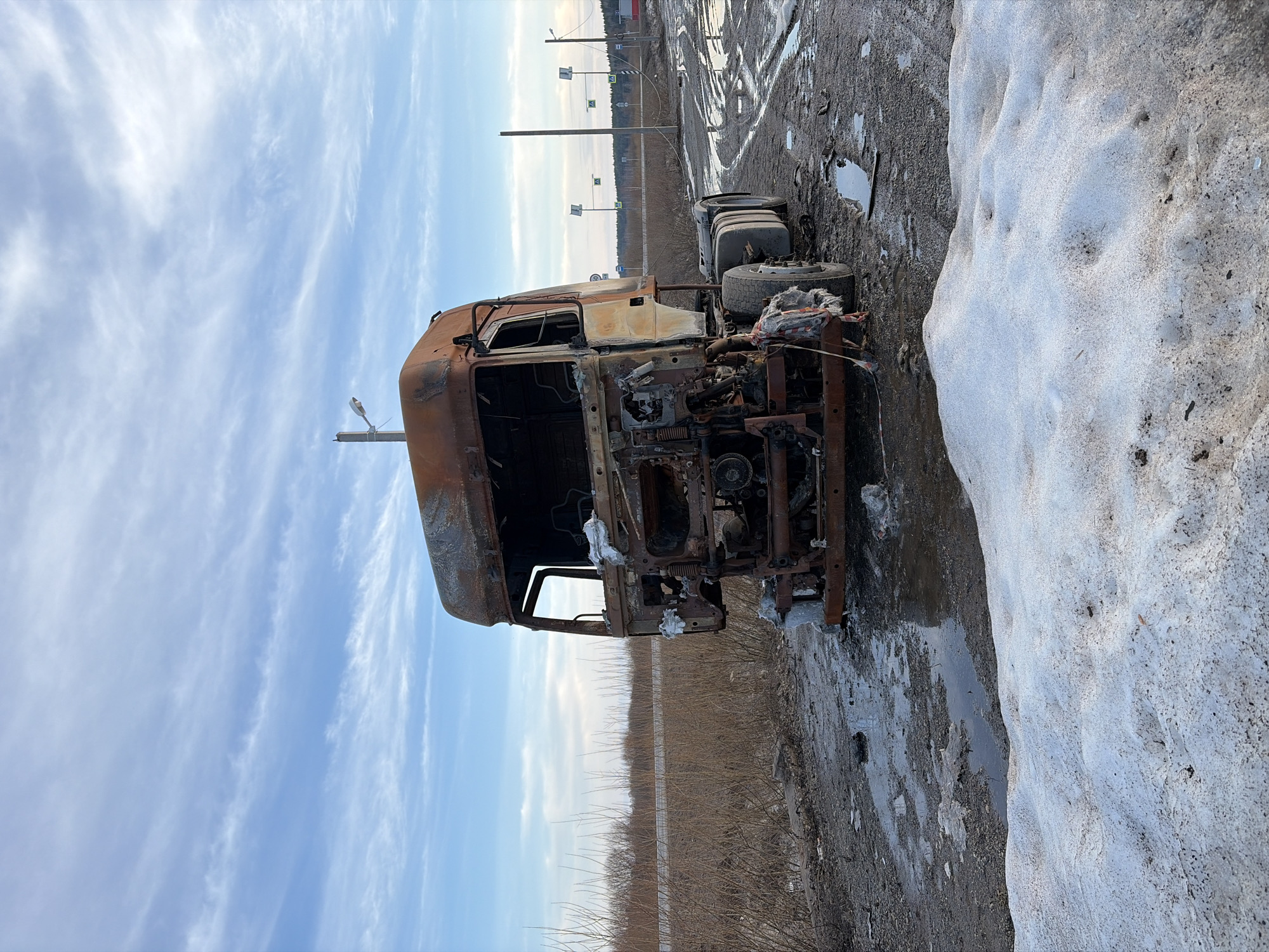 The image shows a severely burned semi-truck cab and chassis in an outdoor environment. The truck is heavily damaged, with most of the cabin structure missing or heavily corroded, indicating a possible fire. The chassis appears to be mostly intact.
The ground is a combination of mud and patchy snow. There is a large patch of snow adjacent to the truck. In the background, poles for streetlights and road signs are visible, along with bare bushes, which suggests the edge of a road or parking area. The sky is lightly clouded.
There is no indication from the image itself that the location is Zachernaya, Russia, but if that information is assumed as correct, then the picture likely depicts an abandoned or burned-out vehicle along the road. No people are visible.