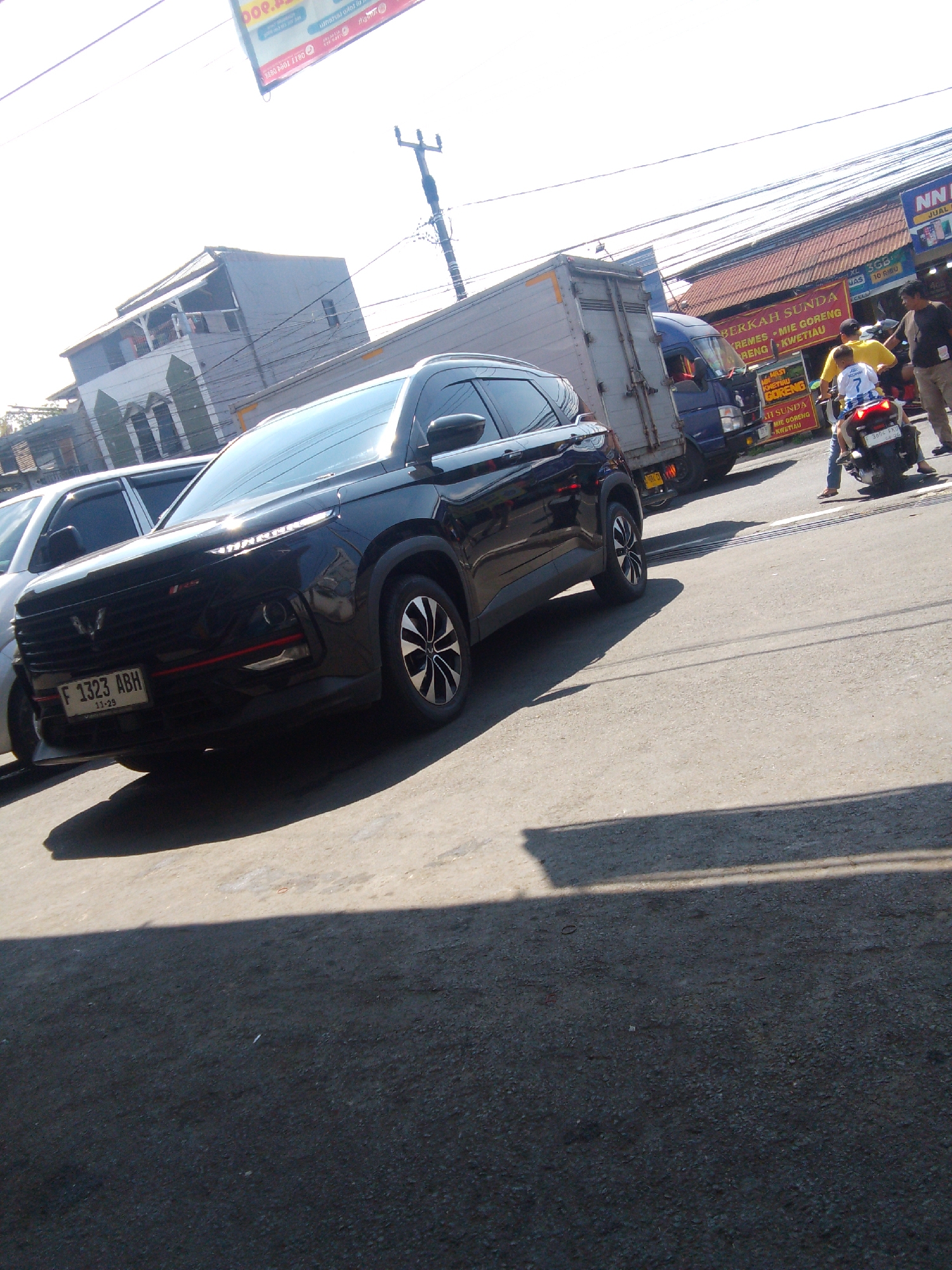 In Cianjur, Indonesia, a black SUV is parked on a street. The vehicle's license plate reads "F 1323 ABH". A white car is parked next to it. In the background, a blue truck is visible, along with several people. One person is riding a motorcycle with a child on the back. A red and white sign is posted on a building in the distance. The sky is bright and sunny.