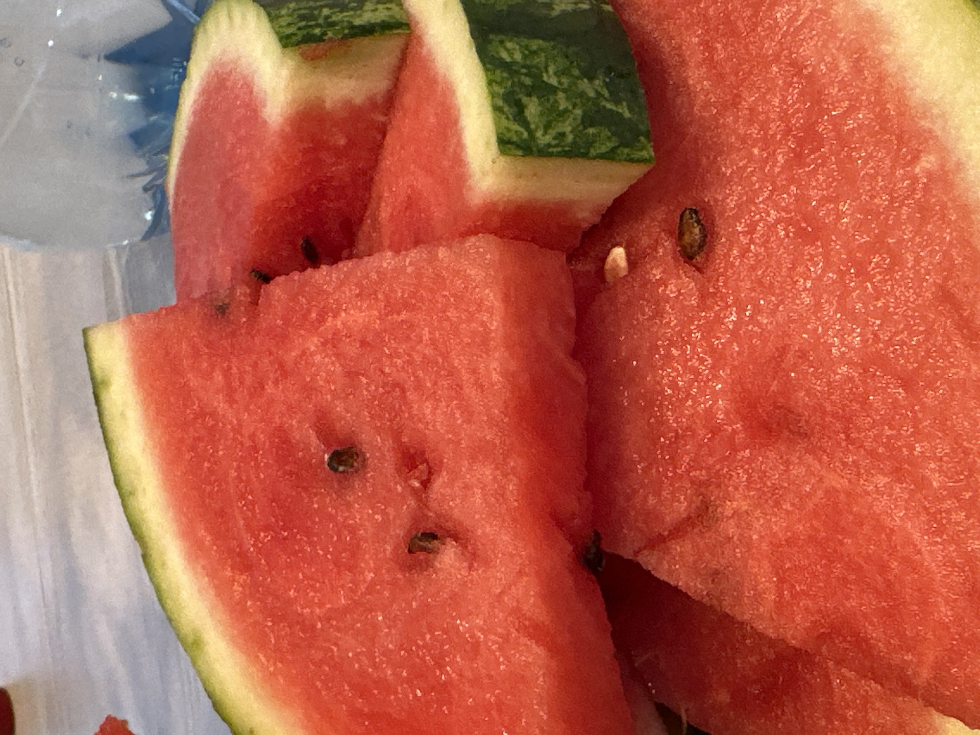 A close-up shot shows several wedges of cut watermelon. The bright red flesh of the fruit, speckled with dark seeds, is prominent. The rind, with its characteristic green stripes and lighter yellow-green layer, is visible on the edges of the wedges. The texture of the watermelon flesh appears juicy and slightly granular. Some of the seeds are visible on the surface and embedded within the flesh. One wedge is positioned prominently in the foreground, with other slices overlapping behind it. The background is blurred and appears to be a light-colored surface, possibly a table or placemat, with a hint of a blue, translucent object which might be a bottle or glass. The lighting suggests it is daytime, and the overall impression is one of freshness and summer refreshment. There is no visible text in the image.