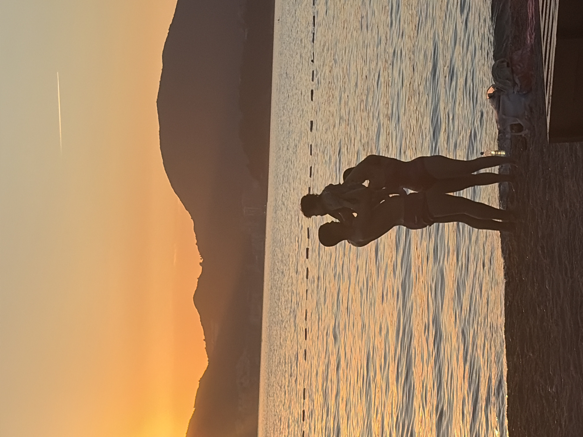 In Sveti Stefan, Montenegro, at sunset, a family is enjoying the beach. A man is holding a baby up in the air, and a woman stands beside him. The sun is setting behind a range of mountains, casting an orange glow over the water. The water is calm with small ripples, and a few buoys mark an area of the sea. The sand on the beach is dark, and there are some items of clothing and a bottle of water near the water's edge. A dark wooden structure is partially visible in the lower right corner of the image. A contrail from an airplane is visible in the sky.