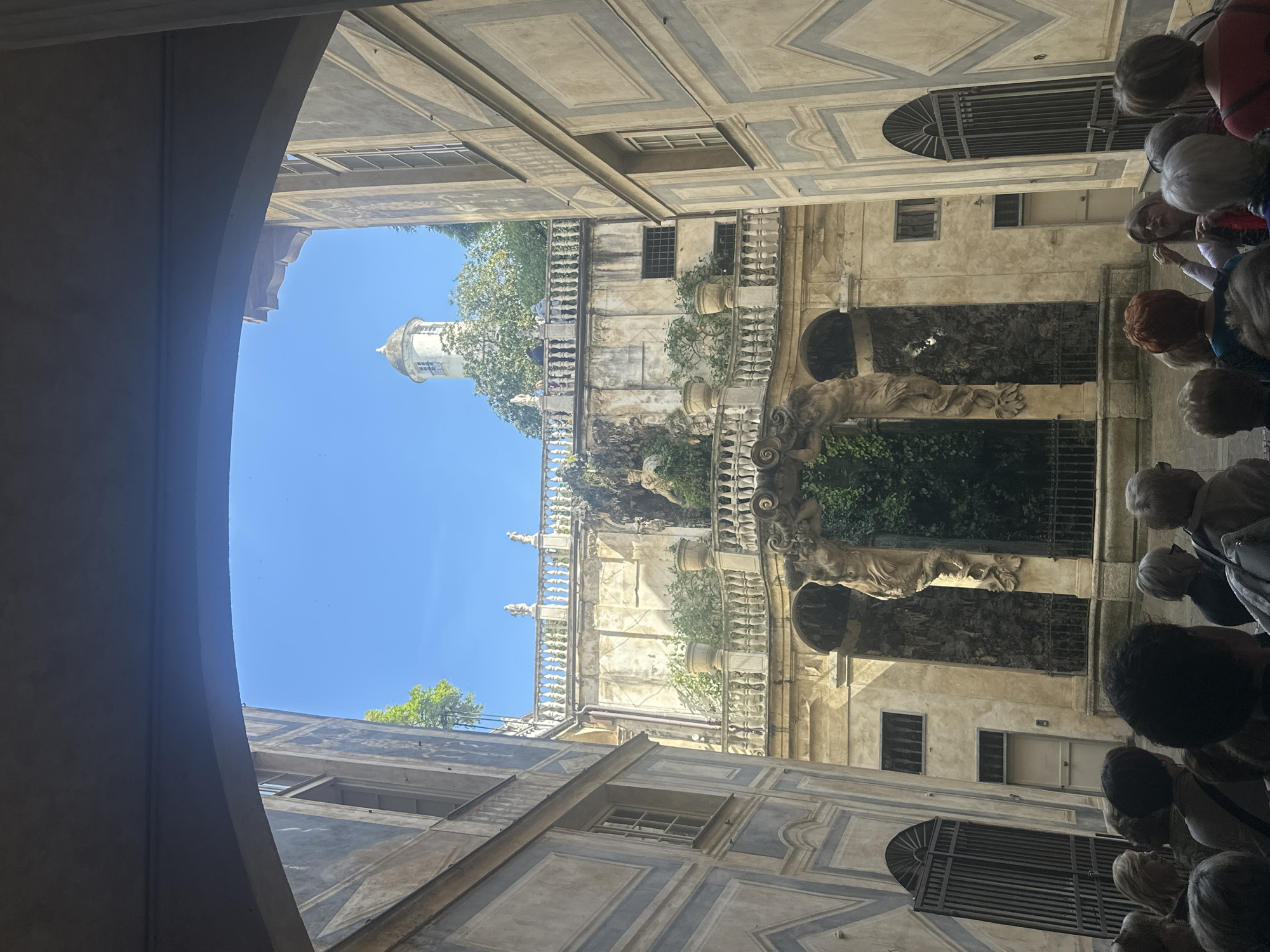 The image presents an upward view from within a shaded, arched passage, looking out into the sunlit courtyard or a grand open space of a historical building complex. The architecture is distinctly classical, featuring ornate facades with painted details resembling coffers and diamond patterns in muted grey and beige tones.
Dominating the central and right portions of the view are multi-story building walls adorned with a series of white, elaborately carved balustrades and balconies. These balconies are interspersed with green foliage, potted plants, and trees, suggesting a verdant garden or terraced landscaping within the complex. Several classical sculptures, depicting human figures and possibly allegorical scenes, are integrated into the balustrade design and recessed wall sections, adding to the grandeur. One prominent sculpture shows a reclining figure. Below these, some wall sections feature dark, textured reliefs, possibly part of a grotto or fountain. Numerous windows of various designs, some with decorative grilles, punctuate the building walls.
Against a clear, bright blue sky in the upper left, a white, multi-tiered bell tower or cupola with arched openings is visible in the distance, partially framed by green tree leaves.
On the far right side of the image, a group of people, likely tourists or visitors, are gathered in the shaded archway, looking out towards the sunlit architectural scene. They are mostly seen from behind or above their heads, dressed in casual attire.
The scene depicts a sunny day with clear weather. The strong contrast between the shade of the archway and the bright sunlight outside suggests it is daytime, possibly morning or early afternoon. The architectural style, with its ornate sculptures, classical elements, and painted facades, is characteristic of historical Italian architecture, consistent with the location context of Genoa, Italy. No visible text is present in the image.