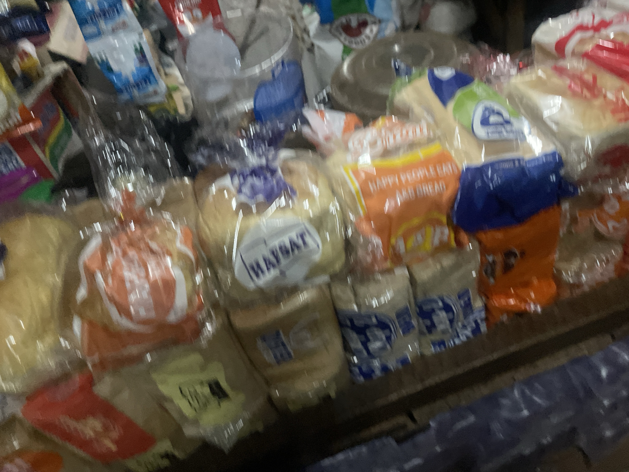 The image depicts a close-up, slightly blurred view of a market stall in Sauka, Nigeria. The stall is laden with various food items, primarily packaged loaves of bread and bottled drinks. The bread packages display different brands and colors, including orange, white, and blue, with some text visible such as "HAPPY PEOPLE EATS" and "MAYSAT". There are also several clear plastic bottles, some appearing to be water bottles. The background is cluttered with more merchandise, suggesting a busy retail environment. The lighting is somewhat dim, contributing to the overall blurriness of the image. No people are clearly visible in the frame.