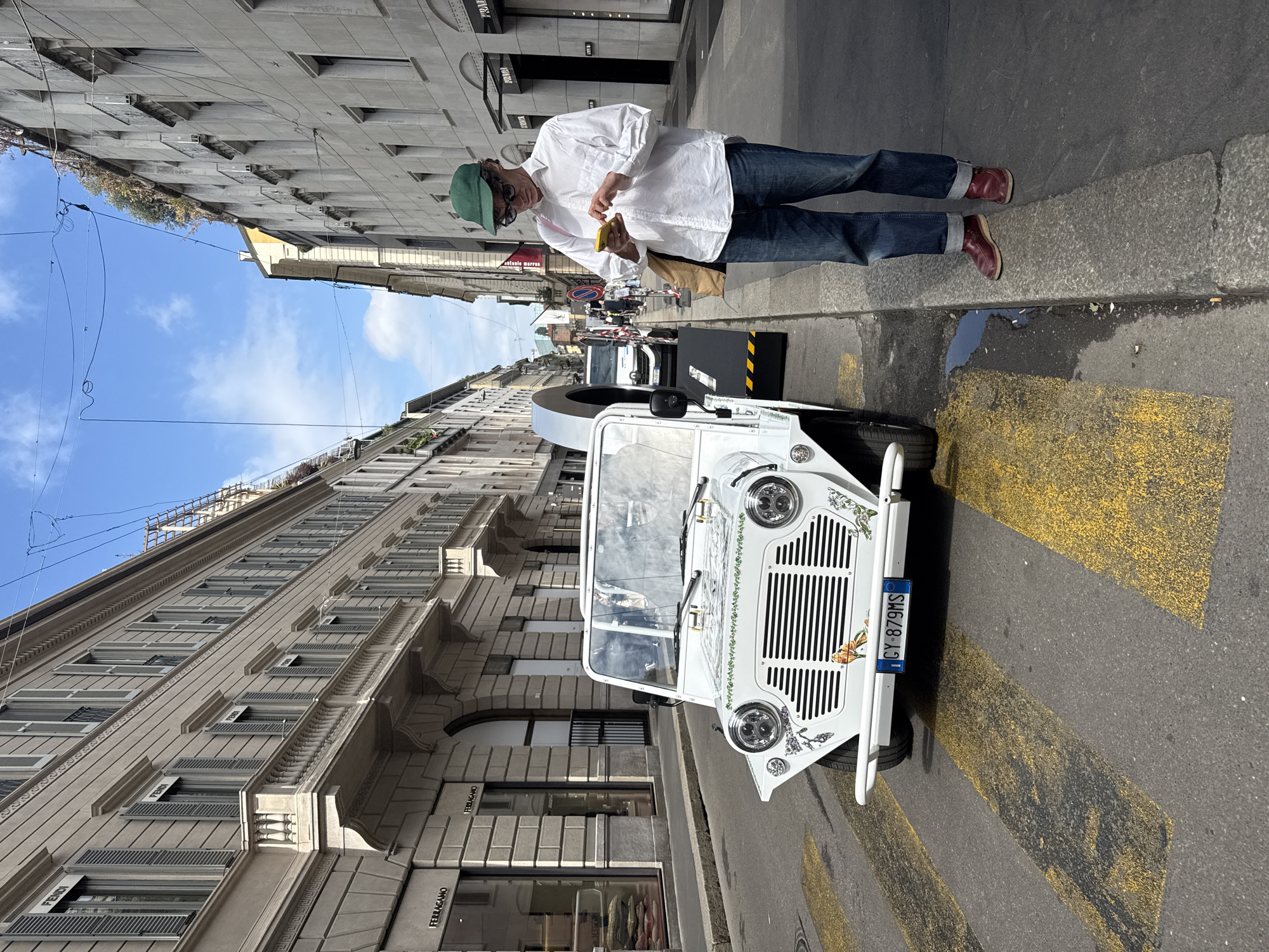 A daytime street scene in Milan, Italy, features a man standing on the sidewalk next to a small, white vehicle parked on the street near a crosswalk. The man is wearing a white button-down shirt, blue jeans, a green cap, sunglasses, and red shoes. He is looking at a yellow object in his hands, possibly a phone or food. The vehicle is adorned with floral decorations and has a license plate that reads "CY-879MS." Buildings with multiple windows and architectural details line the street. The building to the right has Fendi and Ferragamo signs visible. The sky is blue with some clouds visible. The street appears to be clean, with a few wet spots near the curb. There are other indistinct figures and vehicles further down the street, suggesting a busy urban environment.