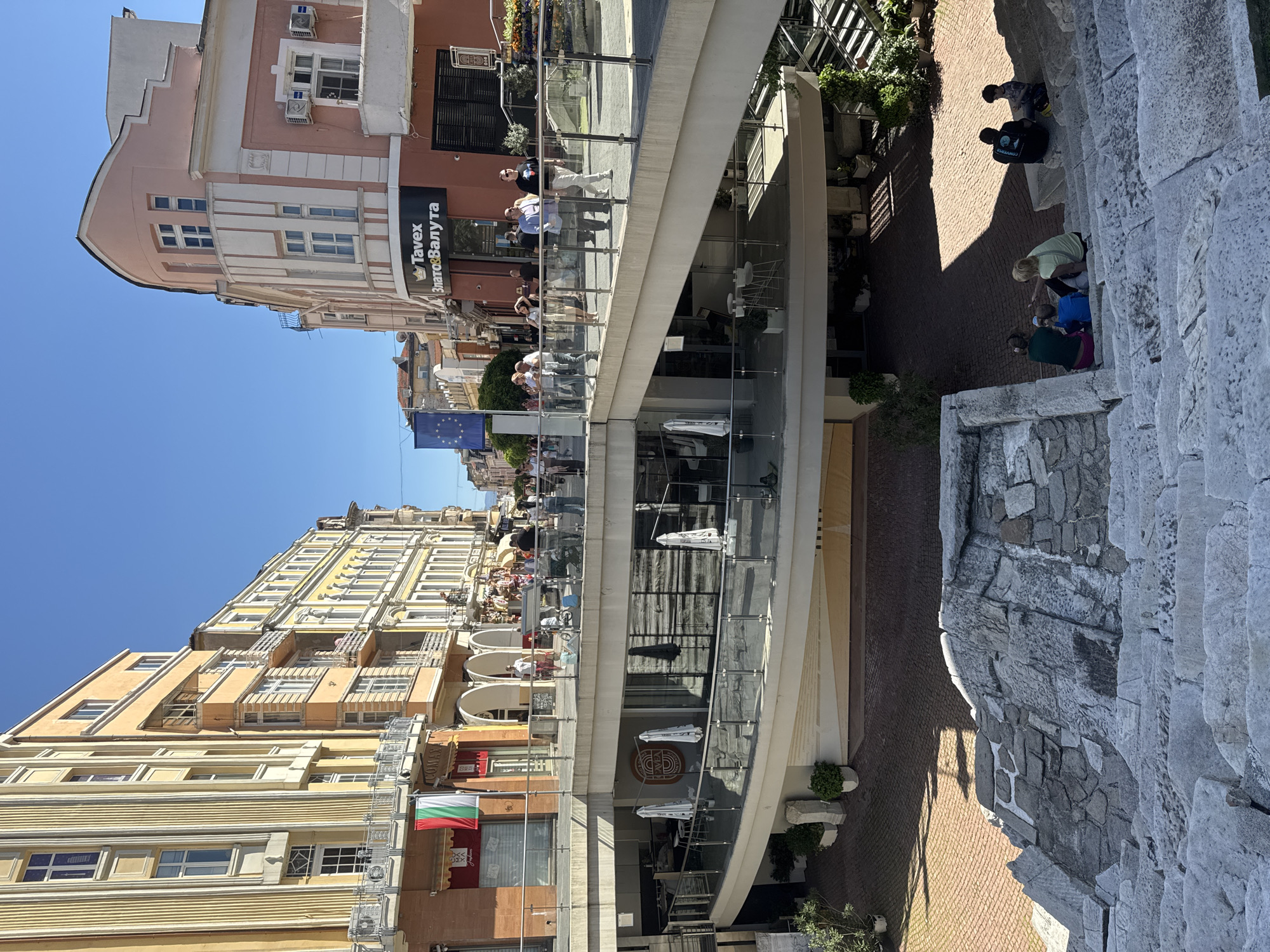 The image shows a street scene in Plovdiv, Bulgaria. On the left is an old-style yellow building with white-trimmed windows. Hanging from the building is the Bulgarian flag. Next to it is a modern building with a curving, glass-paneled walkway extending outward. People are standing on the walkway and looking over the side. On the right of the image, there are Roman ruins with stairs. People are sitting on the ruins and looking around. The sky is a clear, bright blue.