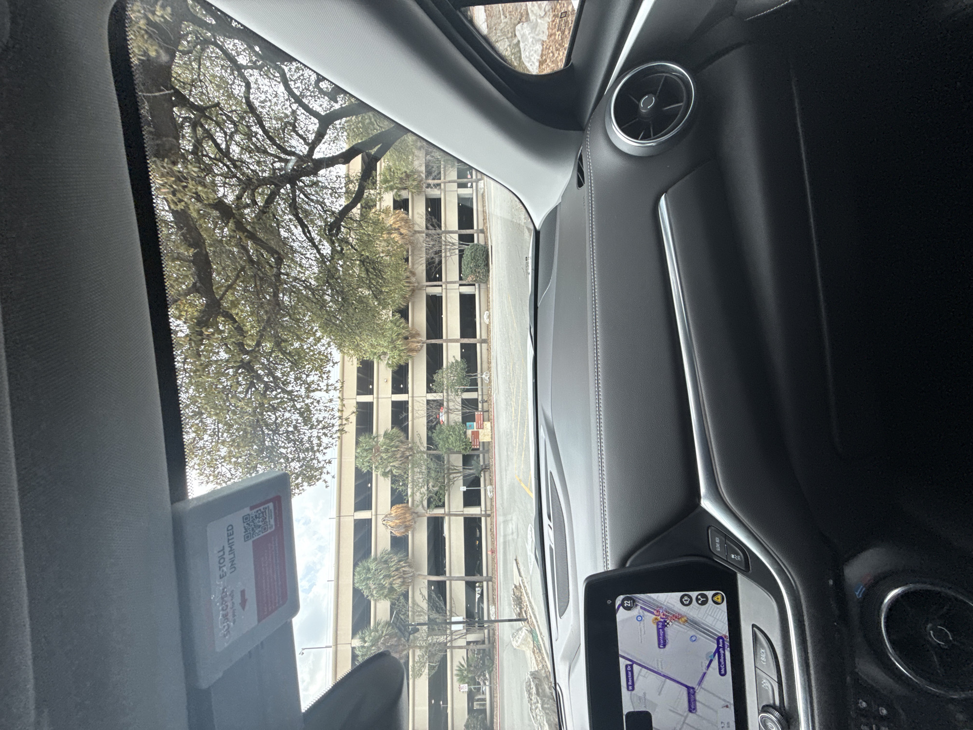 The image, taken from inside a car, shows a view through the windshield of a parking garage in San Antonio, United States. The parking garage is a multi-story building with a light-colored facade and evenly spaced windows. Palm trees and other greenery are visible in front of the garage. Inside the car, part of the dashboard, a navigation screen, and an E-toll device affixed to the windshield are visible.