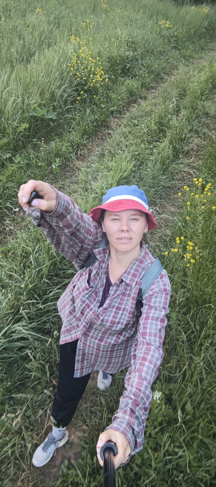 A woman is taking a selfie using a selfie stick in a grassy, outdoor area. She is wearing a red, white, and blue bucket hat, a red and grey plaid button-down shirt over a dark t-shirt, black pants, and grey sneakers. She is also wearing a backpack. The background is filled with tall green grass and small yellow wildflowers. The path appears to be unpaved and winds through the vegetation. The lighting suggests it is daytime, possibly late afternoon or early evening. The image is taken from a high angle, looking down at the woman and her surroundings. There are no other people visible in the image. The location is identified as Garevaya, Russia.
