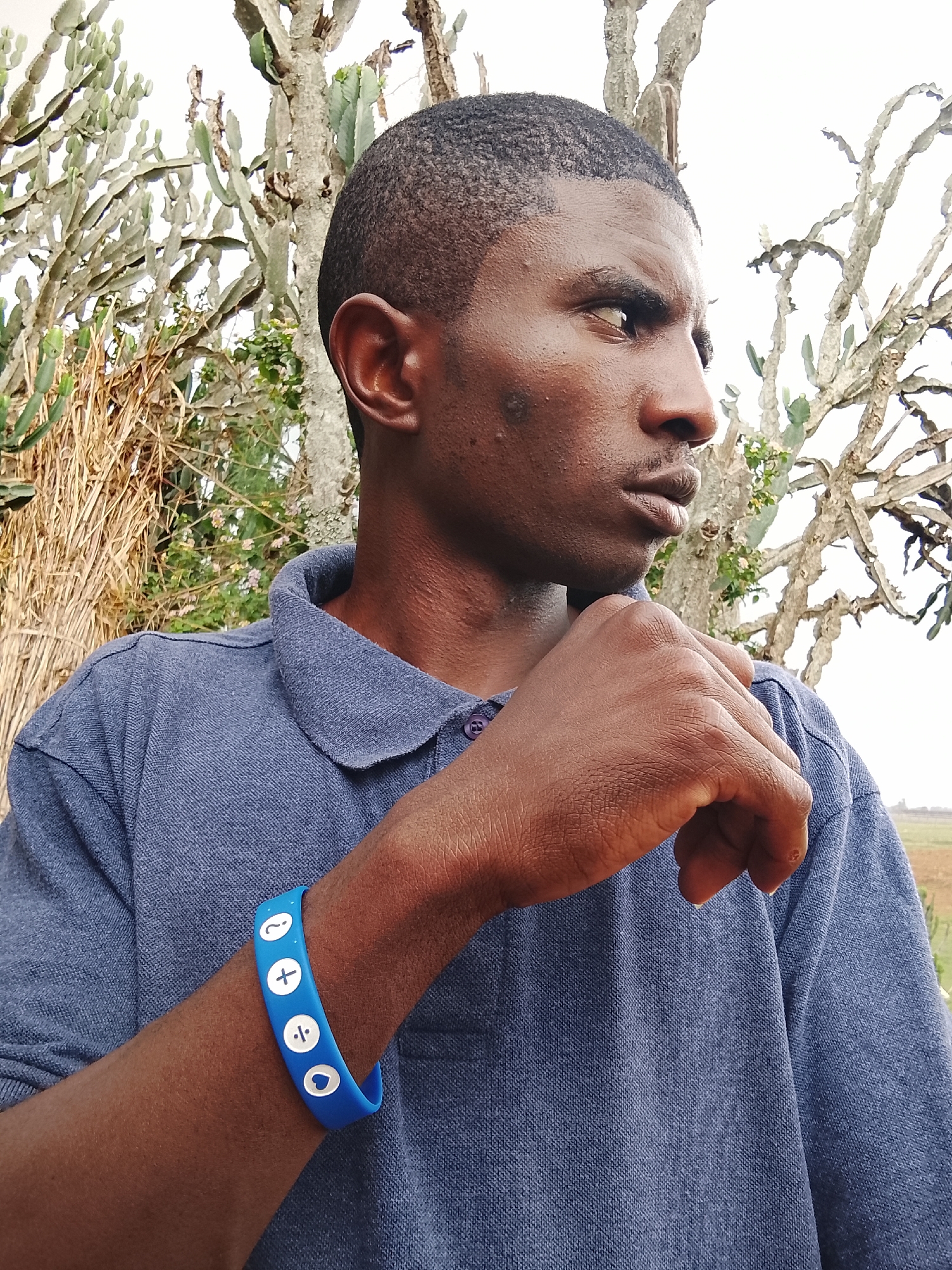 A medium shot depicts a dark-skinned man with short, dark hair wearing a blue collared shirt and a blue wristband with white symbols. His right fist is near his chin, and he is looking to his right with a slightly furrowed brow. Behind him, there are cacti and other vegetation. The location is identified as Unknown, Unknown.