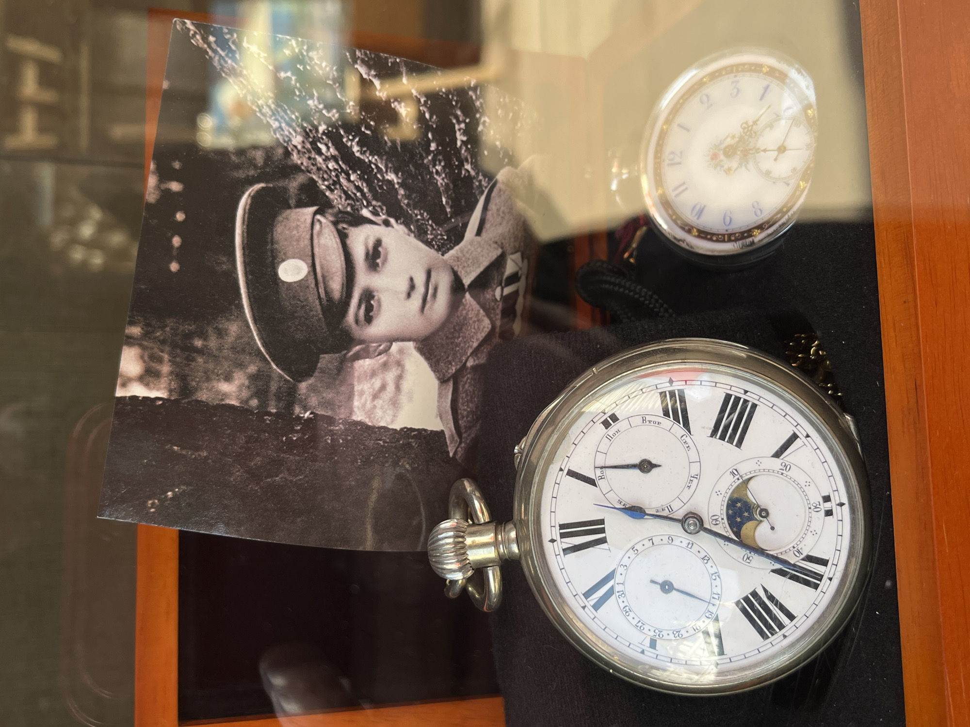 The image captures a display, seemingly within a shop or museum, featuring a collection of vintage items behind glass. On the left, there's a black and white photograph of a young person, possibly in a military uniform, with a serious expression. To the right, there are two pocket watches on a black surface. The larger pocket watch has a white face with Roman numerals and several subdials. The second smaller pocket watch has a delicate design with floral accents. The scene suggests an exhibit showcasing historical artifacts, potentially related to a specific time period or individual. The pocket watch is in Russian and labels the days of the week and days of the month. Based on this information, it is possible that the image was taken in Moscow, Russia.