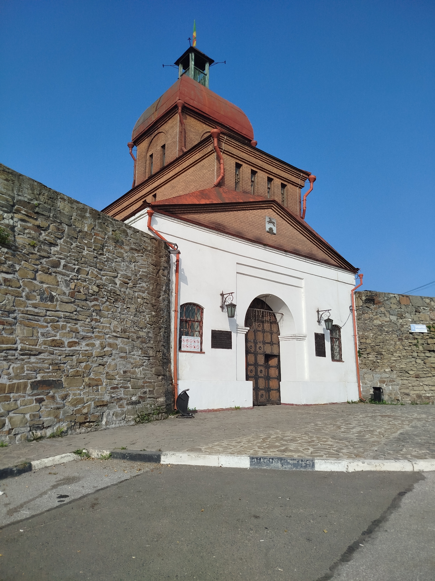 The image shows a view of the Novokuznetsk Fortress (Novokuznetskaya Krepost) located in Novokuznetsk, Russia. The fortress features a prominent red-domed building constructed of brick, with a smaller bell tower on top. A large, weathered stone wall runs along the left side of the frame, leading to the main structure. The main building has a white facade with an arched entrance, a large wooden gate, and decorative lanterns. Two smaller, barred windows are visible on the white section of the building. The ground in front of the fortress is paved with cobblestones and asphalt. The sky is clear and blue, indicating daytime. There are no people visible in the image. The scene appears to be a historical site, possibly a museum or a preserved landmark.