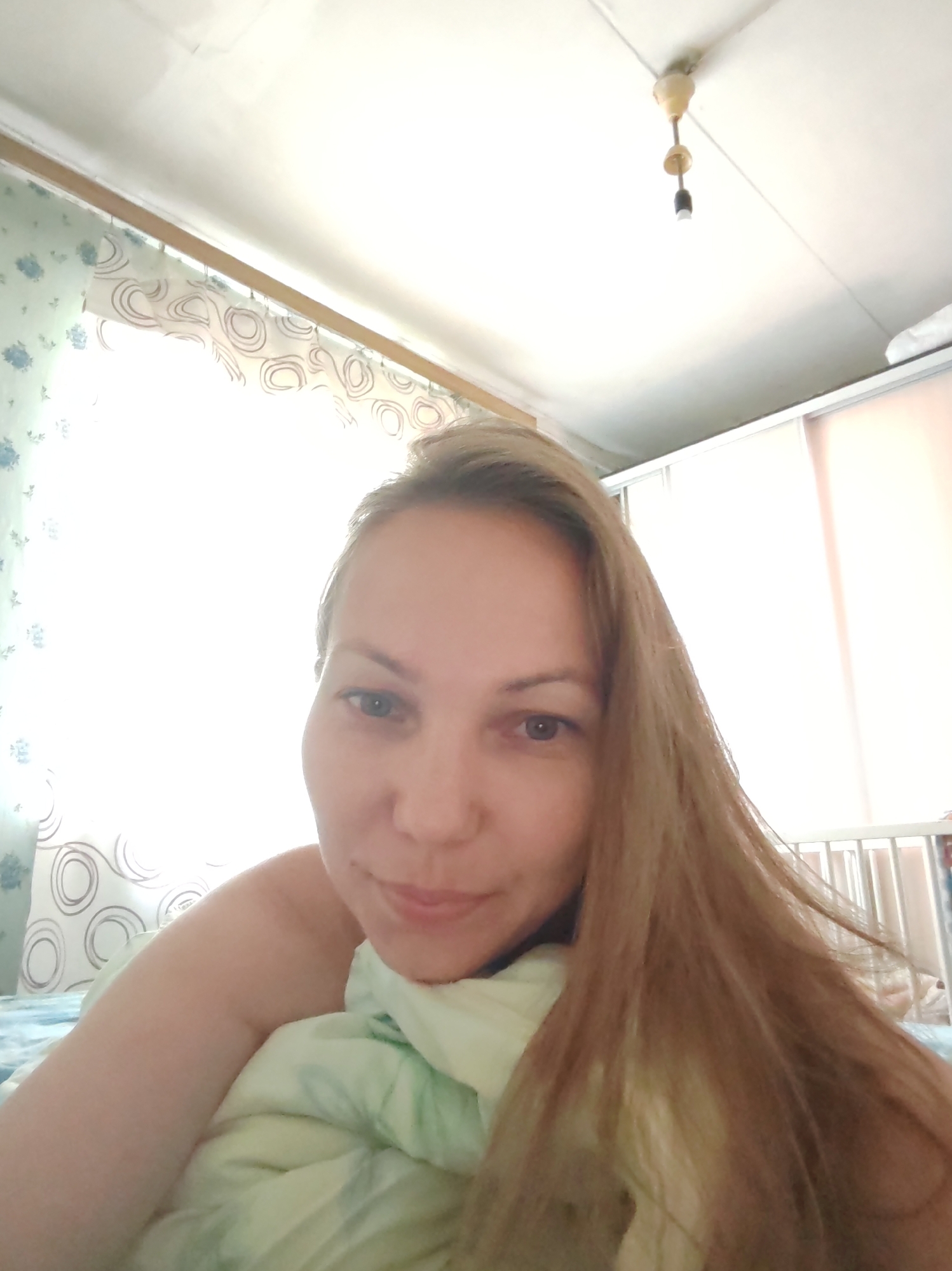 A woman is lying in bed, covered by a light green comforter. She has long, blonde hair and is looking at the camera. The room appears to be a bedroom, with a window in the background. The location is the city of Чухлэм, Russia.