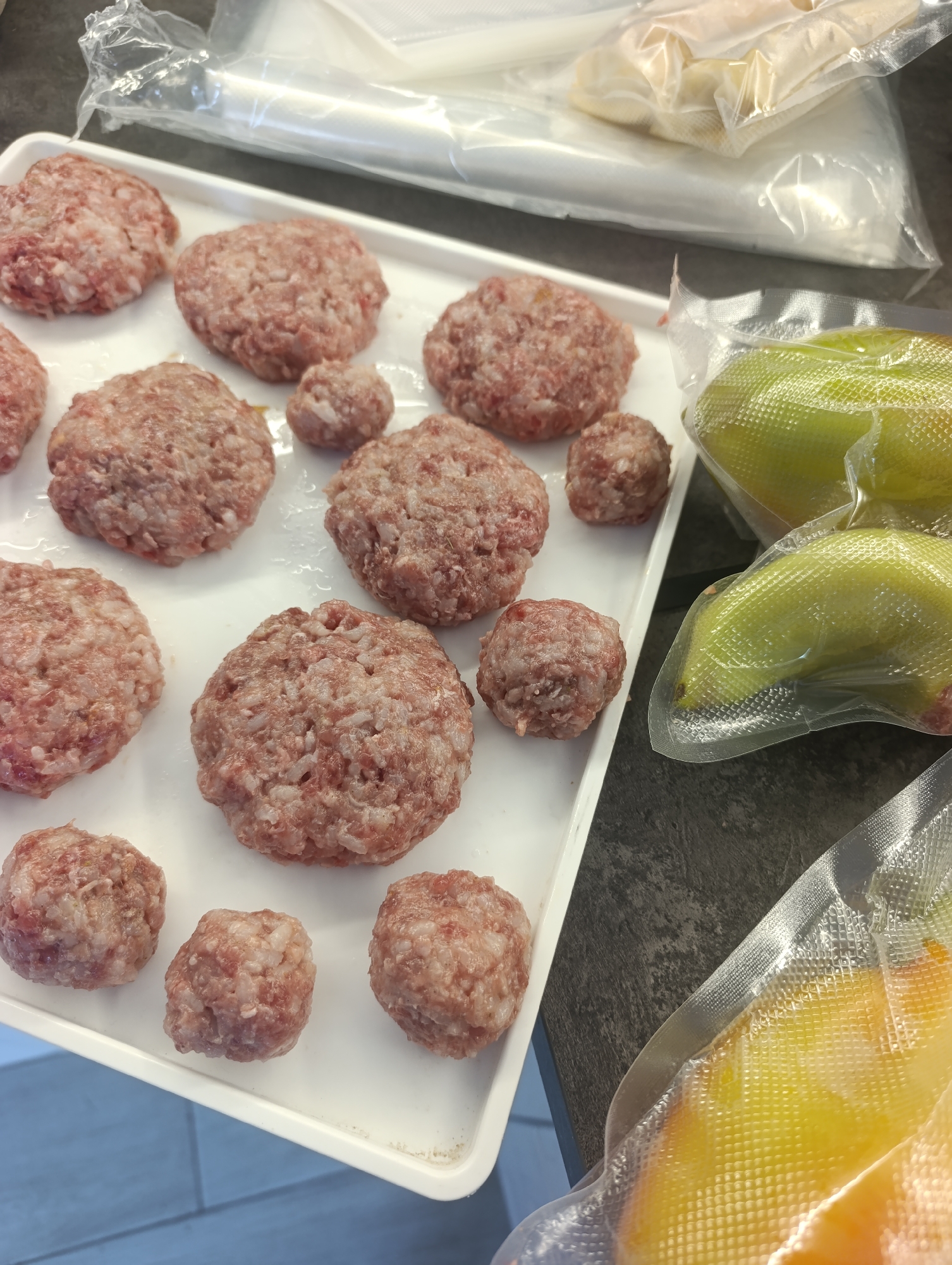 The image depicts a close-up, overhead view of a white tray filled with uncooked hamburger patties and meatballs. The patties are arranged in rows, and some are larger than others. To the right of the tray, there are vacuum-sealed bags containing what appear to be green bell peppers and possibly other produce with yellow and orange hues. In the background, there are more vacuum-sealed bags that are blurred and out of focus. The environment appears to be a kitchen or food preparation area. There are no people or animals visible in the image. The lighting is bright, suggesting an indoor setting. The specific location of Ivanovska, Russia, is not evident from the visual information in the image.