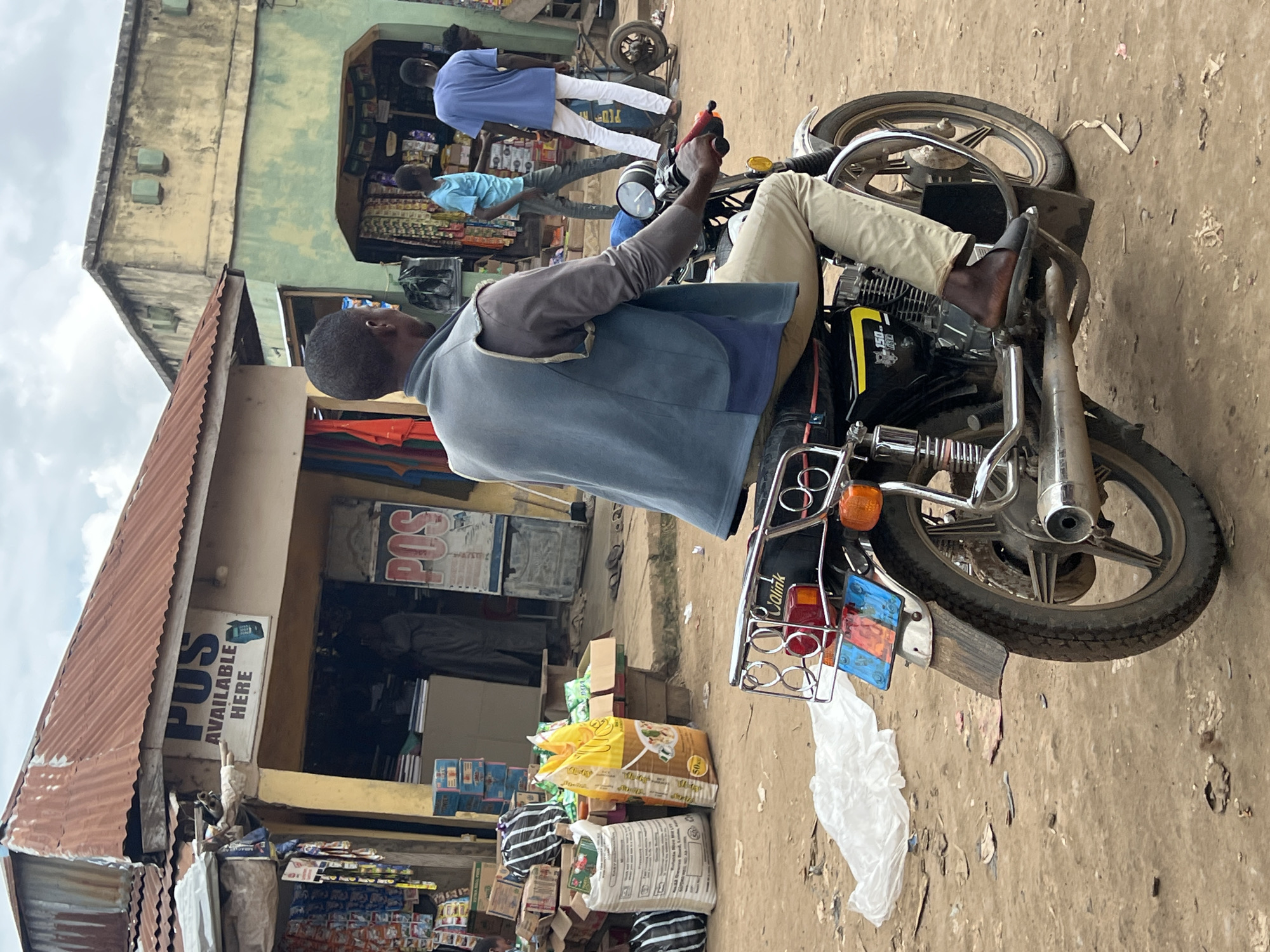 A medium shot captures a man riding a motorcycle on a dirt road in front of shops in Oko Amakom, Nigeria. The man, wearing a light blue vest over a gray long-sleeved shirt and khaki pants, is seen from the back, looking towards the right. The motorcycle is black with yellow accents and a prominent silver exhaust pipe. Behind the rider, a cluster of shops line the street. One shop has a red corrugated iron roof and a sign that reads "POS AVAILABLE HERE" with a graphic of a phone. Another shop, painted green, displays various goods on shelves. Other individuals are visible in the background, including two men in blue shirts, one walking away from the camera and the other interacting with merchandise inside a shop. Stacks of boxed goods and large bags of what appear to be rice are piled near the entrance of one shop. The ground is unpaved and dusty, with scattered debris. The sky is overcast.