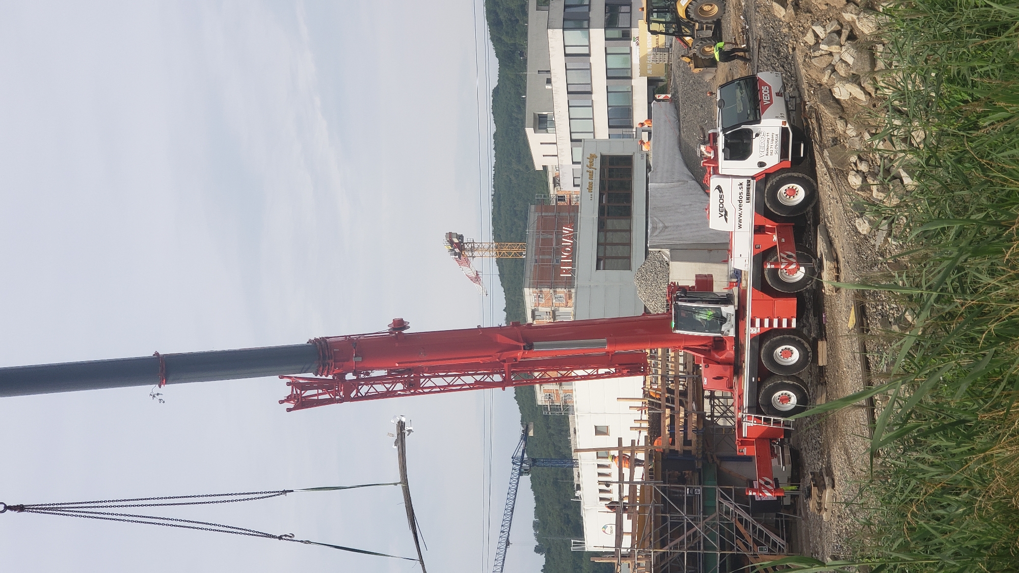 In Prešov, Slovakia, a large red mobile crane, identified by the logo "VEDOS", is actively engaged in a construction project. The crane's boom is extended, lifting a bundle of metal bars. The crane's cab shows a person inside, possibly operating the machinery. In the background, other construction cranes, including a yellow lattice boom crane and a blue tower crane, are visible, along with multiple unfinished buildings under construction. Two construction workers, one wearing a yellow vest, are observed on the ground near the crane and a yellow bulldozer. The foreground of the image is dominated by tall green grass and scattered rocks, suggesting a rugged terrain characteristic of a construction site. The sky is overcast with scattered clouds.