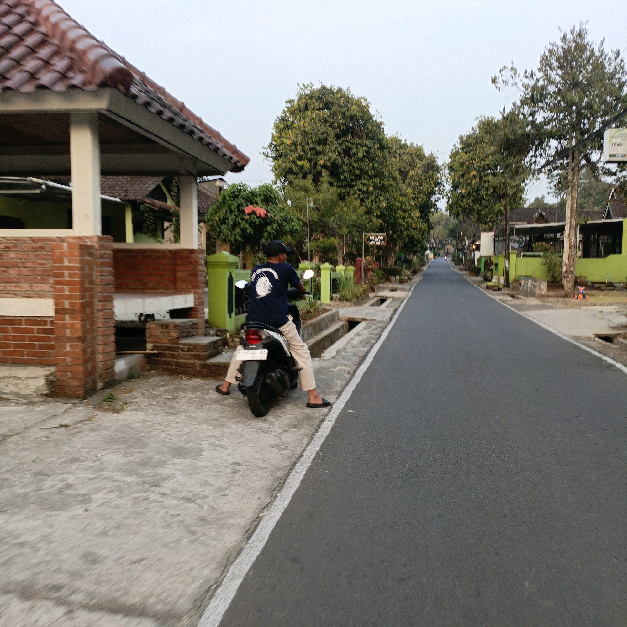 A medium-shot image shows a person on a scooter stopped on the side of a paved road in Salatiga, Indonesia. The person is wearing a dark blue t-shirt with a white graphic, tan pants, a black baseball cap, and dark sandals. They are positioned on the scooter, which is black and white with a license plate. The scooter is on a concrete sidewalk next to a road.
To the left of the scooter is a brick structure with a tiled roof, and a green post. Beyond that, trees and buildings are visible. A sign with text is mounted on a pole in the distance. The road extends into the background, lined with trees and buildings. The sky is overcast. The overall scene depicts a quiet street, likely in a residential or semi-rural area.