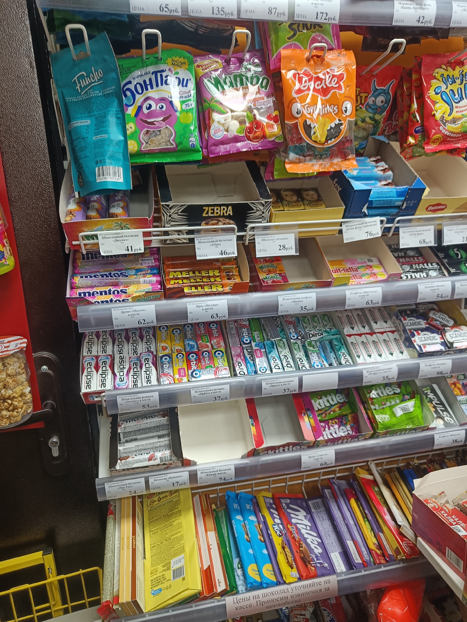 The image shows a shelf in a store in Krasnodar, Russia, filled with various confectionery items. The items are arranged on multiple shelves, with some displayed in cardboard boxes and others hanging from hooks. The products visible include chocolate bars, chewing gum, and candies. Labels and pricing are visible in Russian. The overall environment appears to be a typical retail setting with fluorescent lighting. No people are present in the image.
