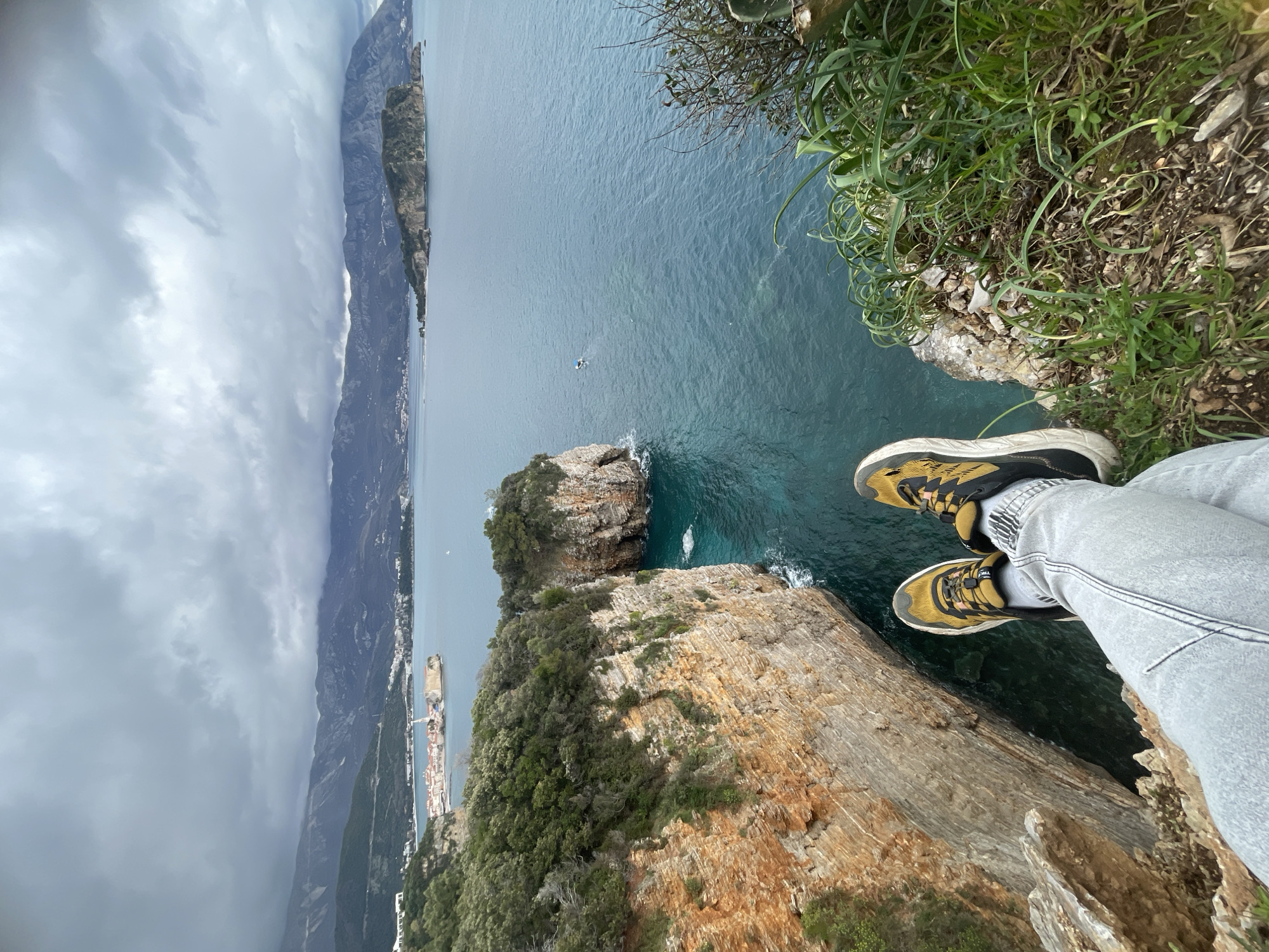 The image depicts a first-person perspective from a clifftop vantage point overlooking the Adriatic Sea, specifically located in Budva, Montenegro. The legs of an individual, wearing light grey jeans and dark yellow and black athletic shoes, are extended over the edge of a rugged cliff.
The immediate foreground consists of sparse green grass and rocky ground at the cliff's edge. Below and to the left, the sheer, stratified rock face of the cliff descends steeply into the turquoise and deep blue waters of the sea. Further out in the midground, the sea extends, exhibiting a rippled surface, with a small white boat visible in the distance.
In the background, the Budva Old Town peninsula is identifiable to the left, featuring historic buildings with red-tiled roofs and a prominent church spire. To the right, Sveti Nikola Island (St. Nicholas Island), a larger, forested landmass, is situated in the bay. A vast range of grey mountains stretches across the horizon, bordering the coastline. The sky is heavily overcast with dense, grey clouds.