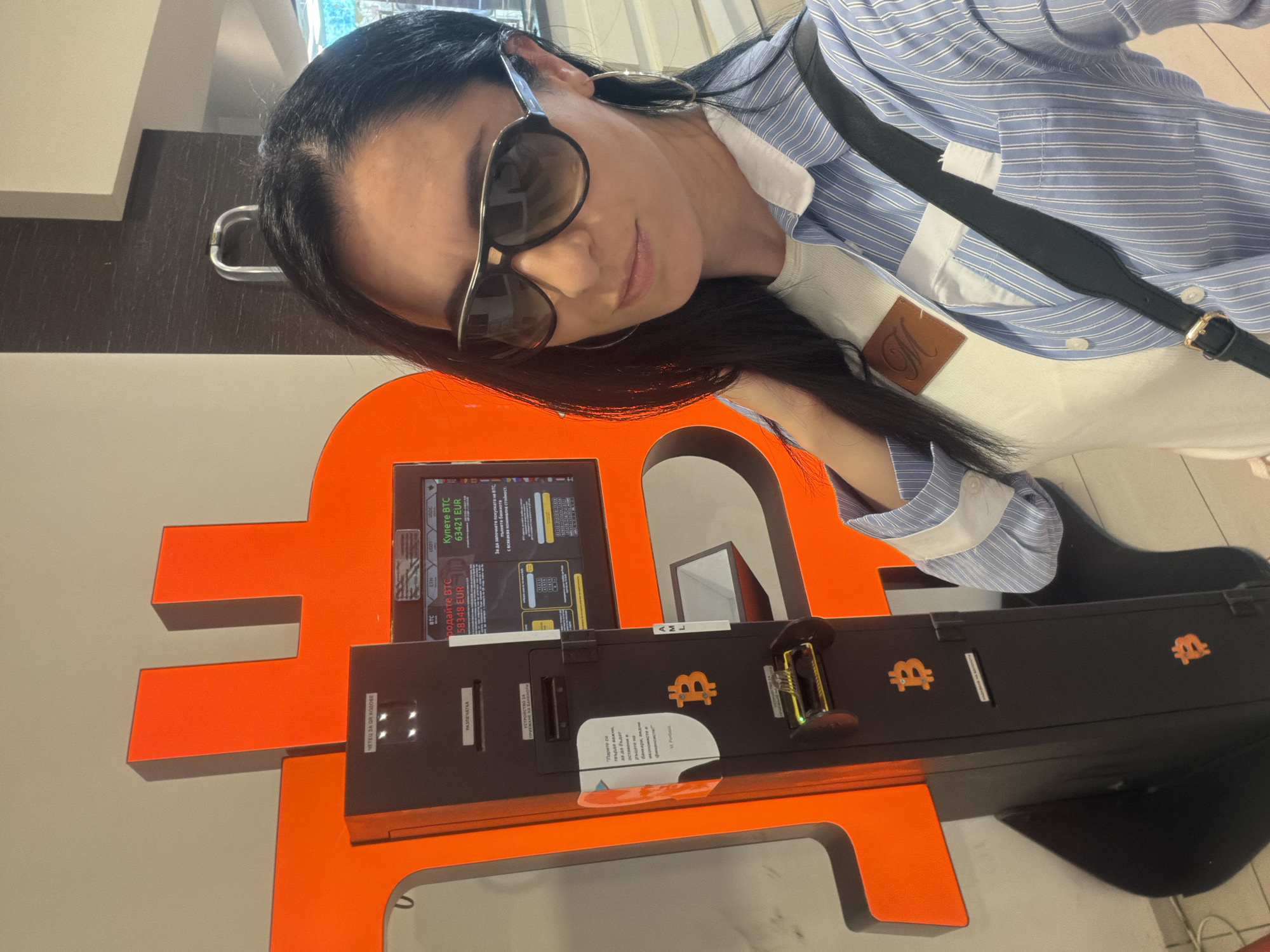 A woman with dark hair and sunglasses is posing next to a Bitcoin ATM. The ATM is primarily black with orange accents in the shape of the Bitcoin symbol. It features a screen displaying information in Cyrillic script, likely related to Bitcoin transactions. The machine has a reader for QR codes and provides an option for printing receipts. The woman is wearing a striped blue shirt and a white top with a brown leather label on the front. A black strap is visible across her shoulder, possibly from a bag. Based on the screen text, the price of Bitcoin for buying and selling is displayed. Additional text on the machine is also in Cyrillic, including information about money and banking. The location is suggested by the text on the machine to be in Bulgaria. Without specific identifying signs or nearby landmarks, a precise location within Pleven, Bulgaria, cannot be determined.