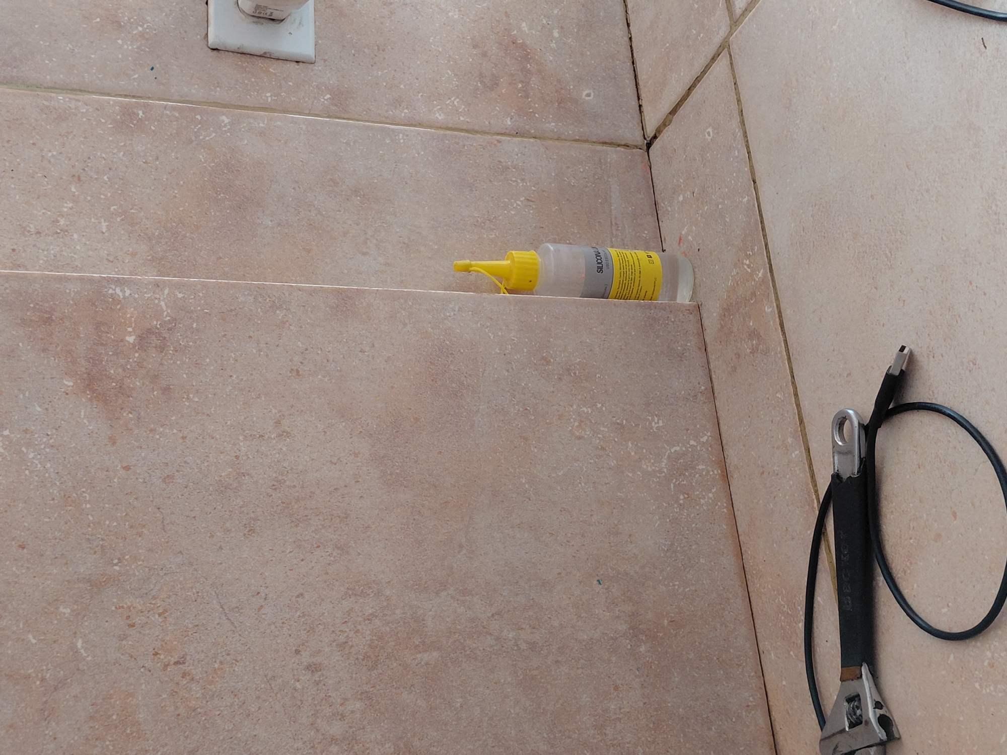 The image captures an indoor scene dominated by light beige or brown speckled tiles, which cover both horizontal and vertical surfaces, forming distinct edges and corners. A white electrical outlet is visible in the upper left corner of the frame, mounted on a tiled wall.
On a lower tiled ledge or shelf, a small, translucent plastic bottle with a yellow pointed cap and nozzle is lying horizontally. The bottle features a yellow label with black text, partially visible, which clearly reads "SILICONA LIQ," suggesting it contains liquid silicone, likely a sealant or adhesive.
In the lower right portion of the image, an adjustable wrench or spanner with a metallic head and a black, possibly rubberized, handle is visible. A black cable, which appears to be a USB cable, is coiled around the handle of the wrench. Faint text, including what looks like the brand "IBEC," is embossed on the wrench's handle.
No people are present, and no activity is actively taking place; the items are stationary. The arrangement of tools and sealant suggests a context of maintenance, repair, or construction, possibly in a utility area, bathroom, or kitchen. The lighting is diffused, consistent with an indoor setting, and there are no discernible cues about the time of day or outdoor weather. The scene is situated in Santa Ana, Venezuela.