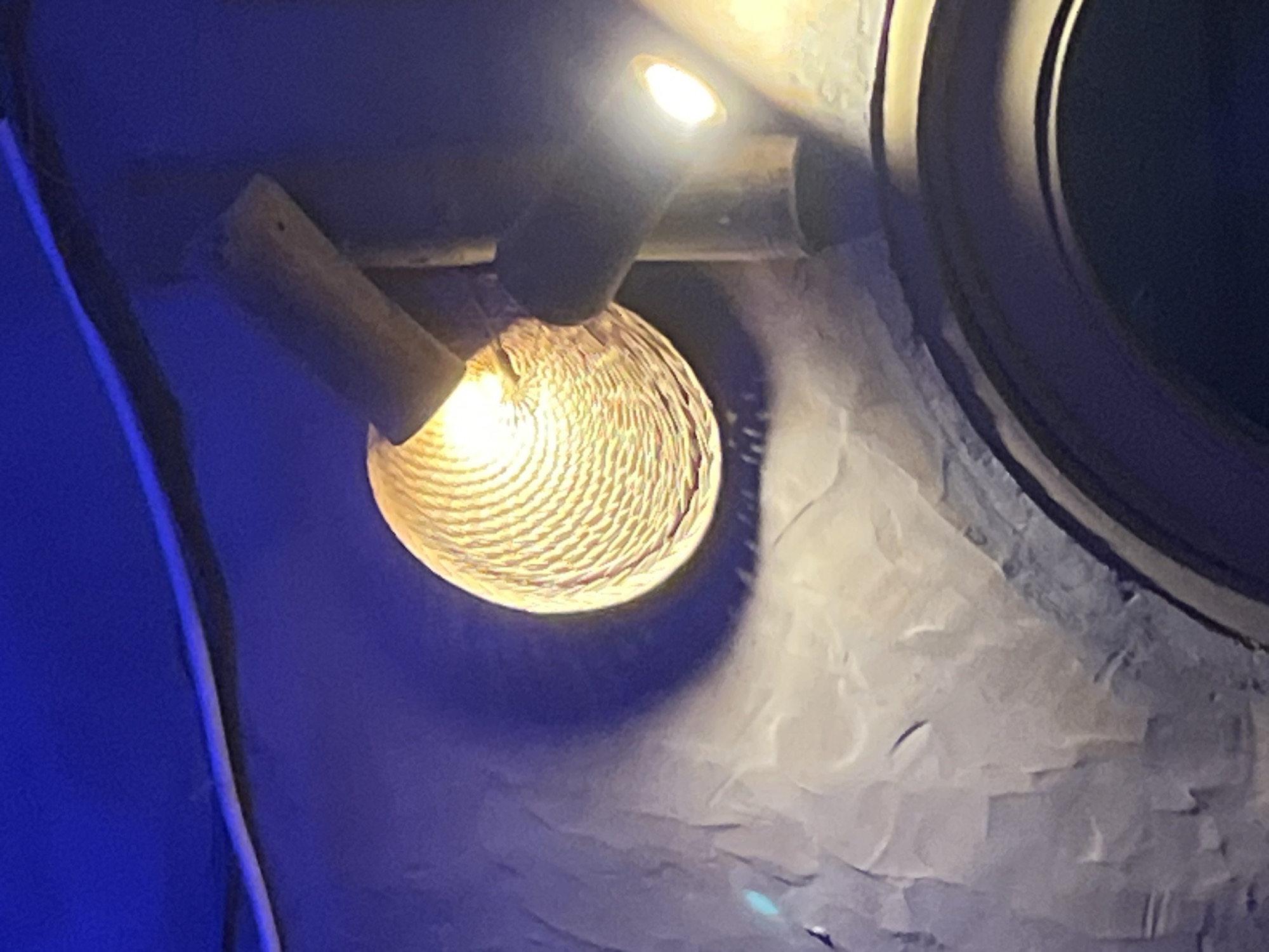 The image shows an interior shot of a building with artistic lighting and textured walls, likely in Mũi Né, Vietnam. A woven lamp with a warm glow hangs from the ceiling, casting shadows on the dark blue walls. The walls appear to be made of rough, uneven plaster or concrete, with subtle variations in color and texture that create a dramatic effect. The lighting suggests a dimly lit, atmospheric space, possibly a restaurant, bar, or a themed attraction. A second light source is visible to the right, also emitting a warm glow. The overall impression is one of rustic elegance and an intimate, possibly bohemian, ambiance.
