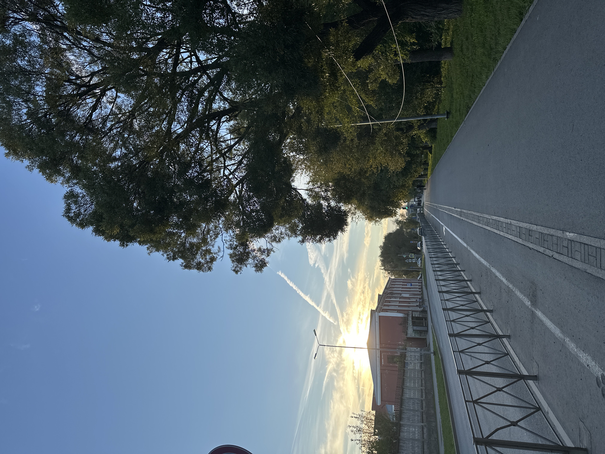 The image depicts a paved path alongside a road in Perm, Russia, bordered by a metal fence on one side and a row of trees on the other. The sun is low in the sky, casting a warm glow and creating long shadows. The sky is blue with scattered white clouds and contrails. There are buildings and vehicles visible in the background. The path appears to be a pedestrian walkway or bicycle lane, separated from the road by the fence. The overall scene is one of a typical urban park or street, with elements of nature and infrastructure coexisting.