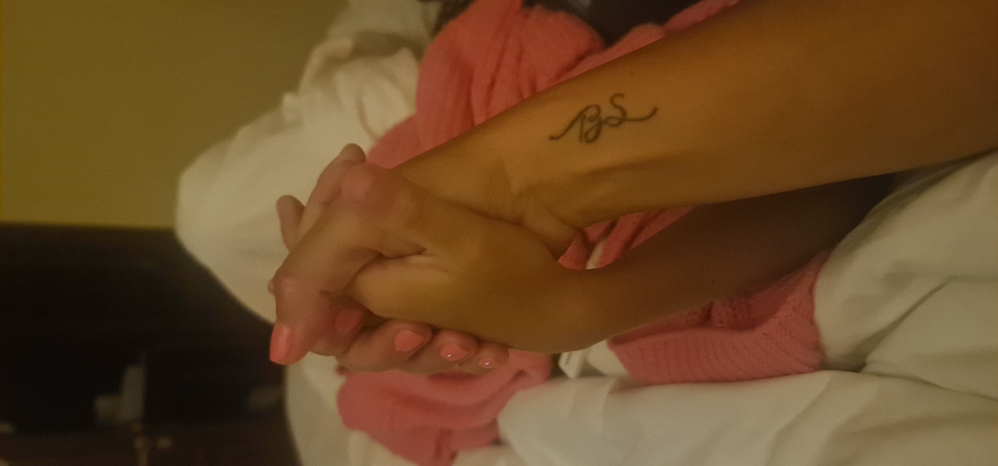 A close-up, medium shot depicts a person's two hands intertwined, resting on a surface with a pink knitted garment and white bedding. The individual has a tattoo of what appears to be the letters "BS" in cursive script on their left forearm. The nails are painted a light pink color. The background suggests an interior setting, potentially a hotel room. The specific location within Đà Nẵng, Vietnam, is not discernible from the image.