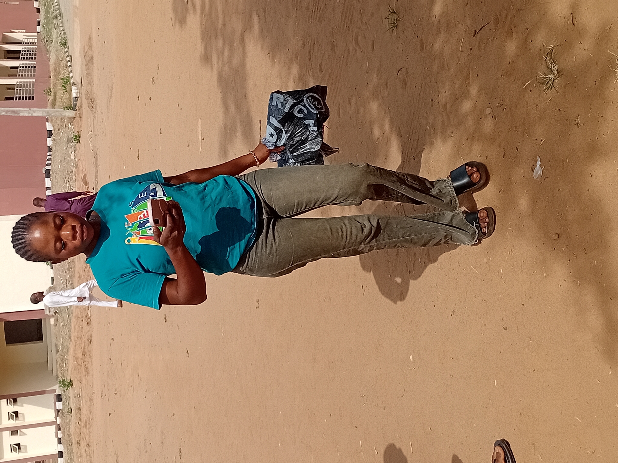A woman stands outdoors in a sandy area, possibly in Lokoja, Nigeria. She wears a blue t-shirt, green pants, and sandals. She holds a phone in her right hand and a black plastic bag in her left. In the background, buildings are visible, as are other people. The environment appears dry with sparse vegetation.