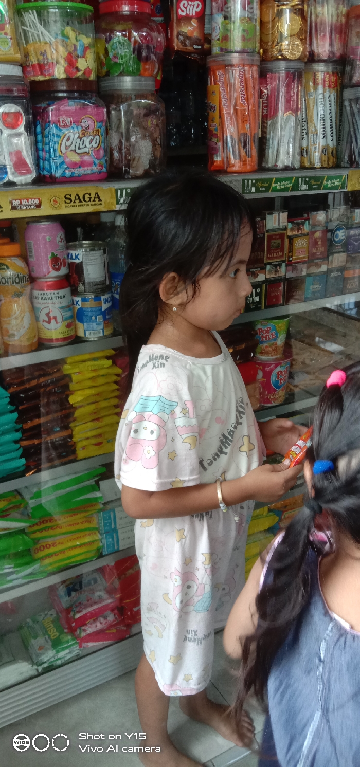 The image depicts an indoor scene within a retail store, identified as being in Sebaung, Indonesia. Two young children are present in the foreground and mid-ground, standing before shelves stocked with various consumer products.
In the mid-ground, a young female child faces towards the left, presenting her right profile. She has dark hair tied back in a ponytail and wears a small earring in her left ear. Her attire consists of a light-colored short-sleeved t-shirt patterned with cartoon characters and text. A gold-toned bracelet is visible on her left wrist. She is barefoot and holds a small, orange-packaged item in her hands, directing her attention forward.
Partially visible in the immediate foreground, with her back to the camera, is another child. This child has dark hair, potentially secured with a blue hair tie, and is wearing a dark blue garment.
The background consists of multi-tiered display shelving filled with merchandise. The top shelves hold transparent plastic jars containing assorted candies and packaged snacks, including "Superstar" wafers and "Candy Choco." Below these, beverages like "Floridina" orange juice, "Larutan Penyegar Cap Kaki Tiga," and canned "Indomilk" are visible, alongside stacks of yellow and green packaged goods, some priced at "Rp 2000." Further in the background, a display unit contains multiple rows of cigarette packs from various brands, including "Gudang Garam," "Sampoerna A Mild," and "Marlboro," with "SAGA" and "SULUH" branding prominent. The floor is light-colored and tiled. The scene is brightly illuminated, characteristic of a small commercial establishment.