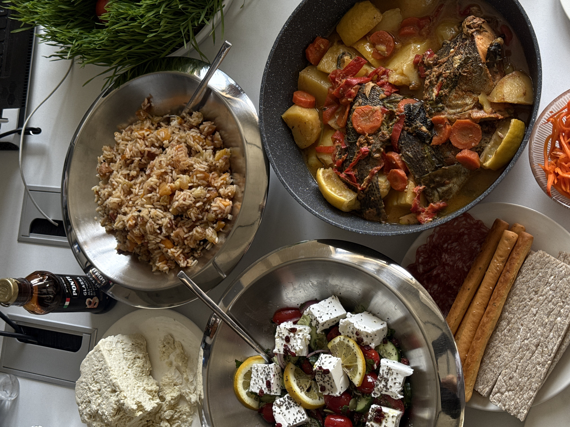 A top-down view reveals a spread of food arranged on a light-colored surface, possibly a countertop or table. To the upper left is a bowl of rice mixed with pieces of fruit, next to a pot containing fish stewed with potatoes, carrots, tomatoes, and lemon wedges. In the lower part of the frame is a bowl of salad with cubed feta cheese, tomatoes, cucumbers, lemon wedges, and seeds, next to a plate of salami, breadsticks, and flatbreads. A bottle of balsamic vinegar and a spread, possibly hummus, are visible near the bottom left corner. The setting appears to be indoors, and the absence of external contextual clues makes it difficult to confirm the specific location. Therefore, it cannot be determined if it is located in Yerevan, Armenia, without further information.