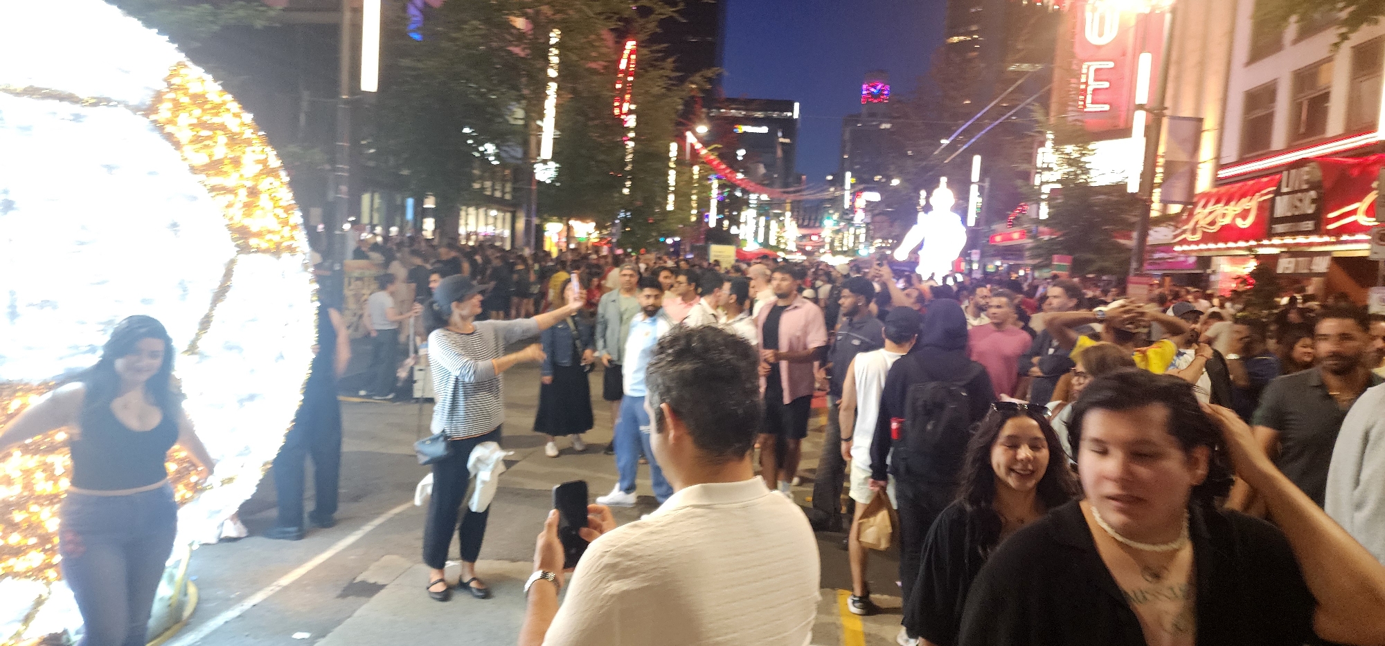 The image captures a bustling street scene at night, likely in a city center. A large, illuminated spherical decoration is prominent on the left side, casting a bright glow. A woman in a black tank top and blue jeans stands in front of it, looking to her left. The street is crowded with people walking and mingling. Buildings with glowing signs and lights line the street, suggesting a commercial or entertainment district. Some signs are illuminated in red and white. The sky is dark, indicating it is nighttime. The overall atmosphere appears lively and festive, with many individuals engaged in various activities, some holding phones, suggesting photography or communication.