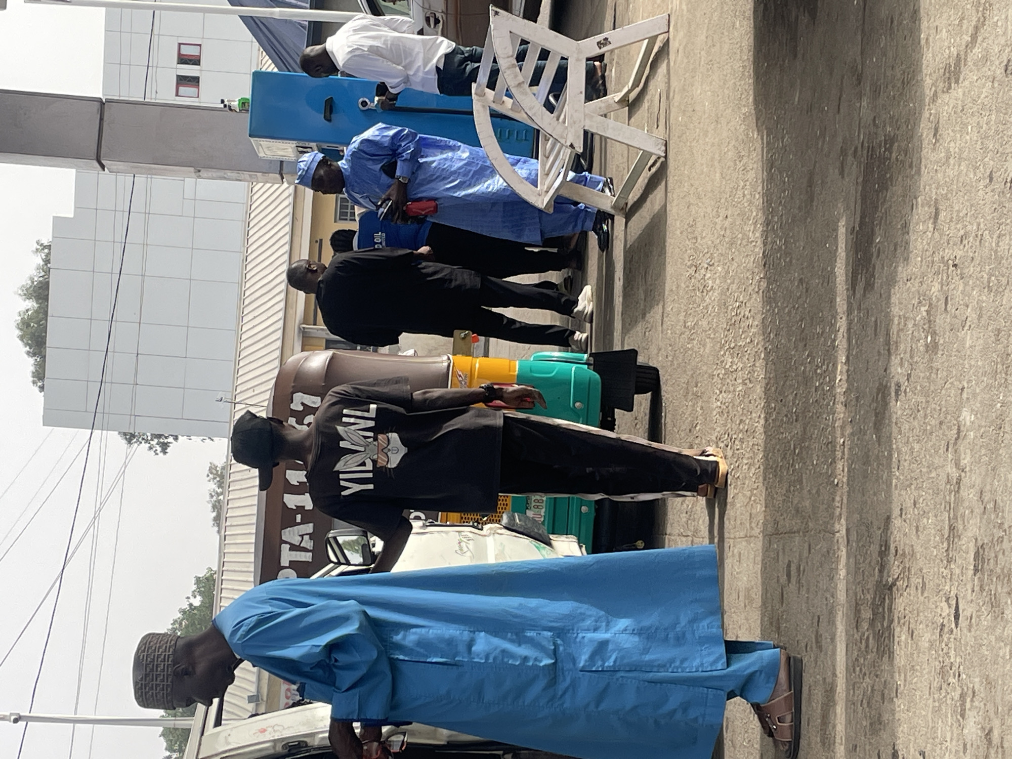 The image captures a street scene in Jos, Nigeria. A line of men stands near a gas pump, potentially waiting to refuel vehicles. The individuals are dressed in a variety of styles, including traditional clothing (flowing robes in shades of blue and black) and casual wear (t-shirts and trousers). A "keke" (a three-wheeled motorized vehicle) is visible among other automobiles. The background shows a modern building and typical city infrastructure. The scene suggests a routine activity in an urban environment, possibly related to fuel consumption.