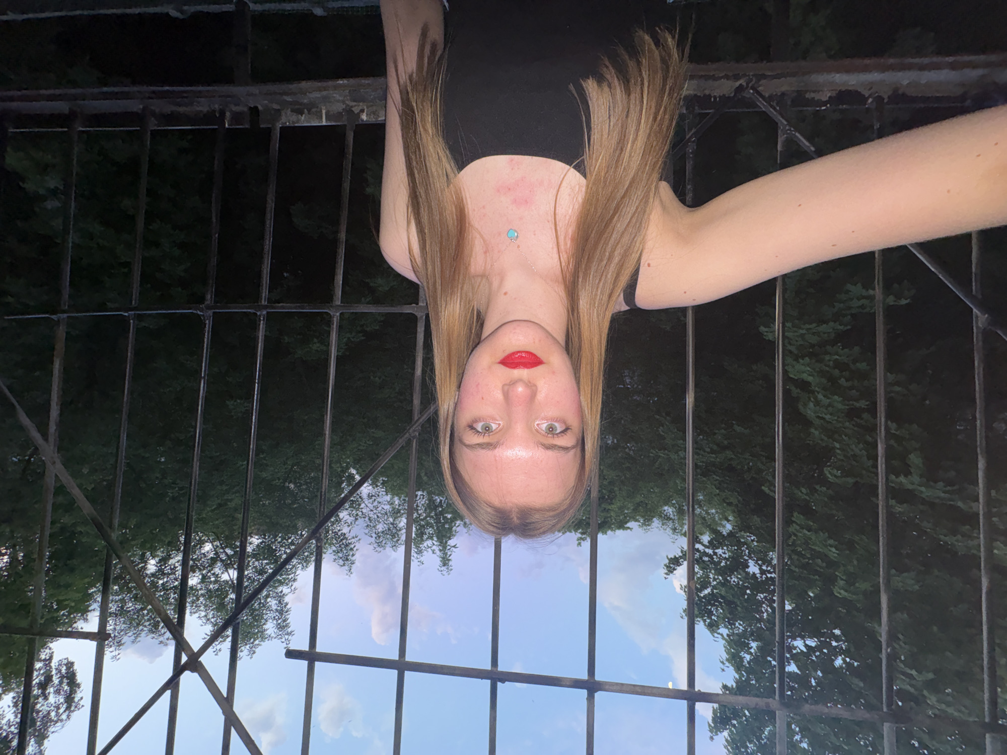 A woman with long, light-brown hair is photographed upside down, visible from the chest up. She is wearing a black tank top and a turquoise necklace. Her lips are painted bright red. She is holding onto a dark metal fence. Behind the fence are trees and a twilight sky with clouds. The location is Perm, Russia.