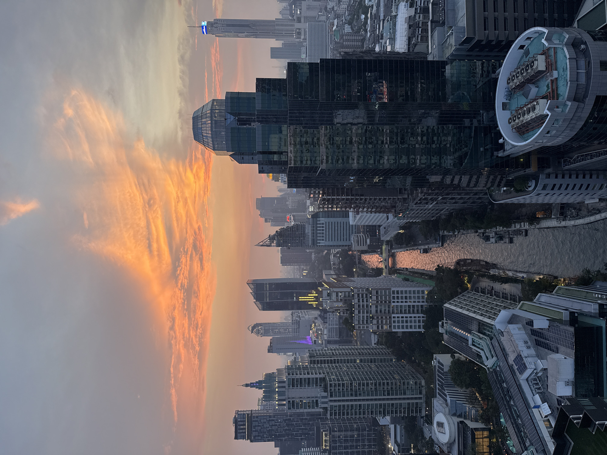 The image depicts a high-angle view of Bangkok, Thailand, during a sunset. Predominantly featuring the city's skyline, the photograph showcases numerous skyscrapers and buildings. A river winds through the urban landscape. The sky is filled with warm hues of orange and yellow from the setting sun. Some buildings have lights on, suggesting it is nearing dusk. Details visible on rooftops include architectural elements and what appears to be ventilation equipment. The general mood is one of urban density and vibrant twilight.
