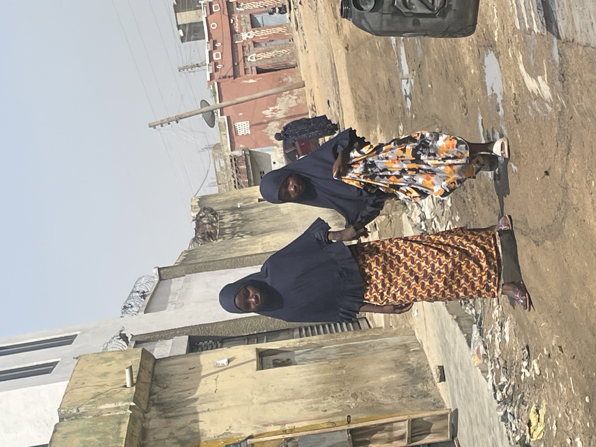 Two women wearing dark-colored head coverings and patterned dresses stand outside a weathered, multi-story building in an urban environment. Overhead power lines run across the sky. Litter is scattered across the ground. Another person is seen in the background near a red building.