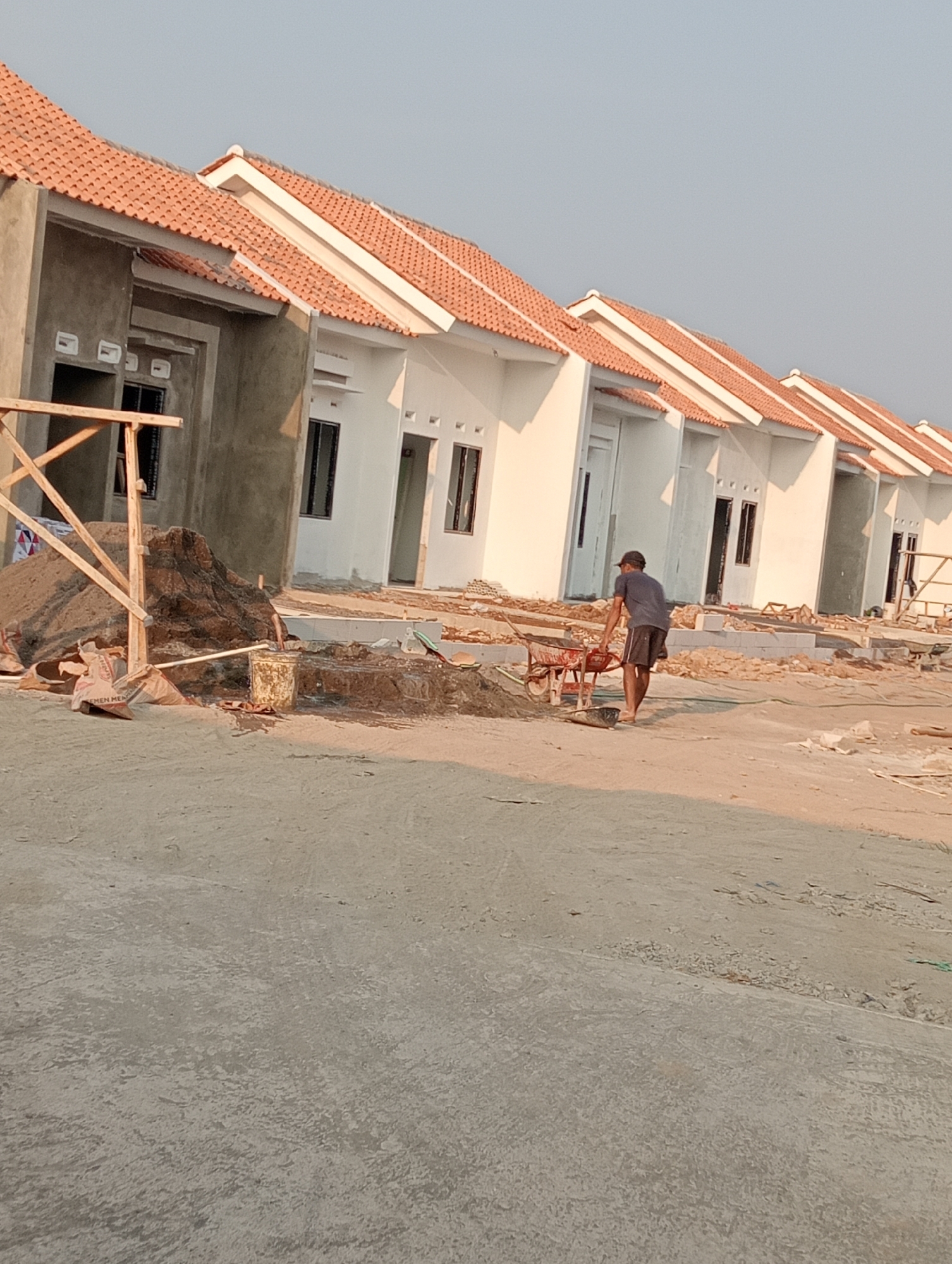 The image depicts a row of houses under construction in the city of Kenep, Indonesia. The houses have white walls and orange tiled roofs. In the foreground, a man wearing a dark t-shirt and shorts is pushing a red wheelbarrow. Piles of sand and construction materials are scattered around the site. The ground appears to be dirt and concrete. The sky is clear and a warm, golden light suggests it is either early morning or late afternoon. The overall scene is one of active construction.