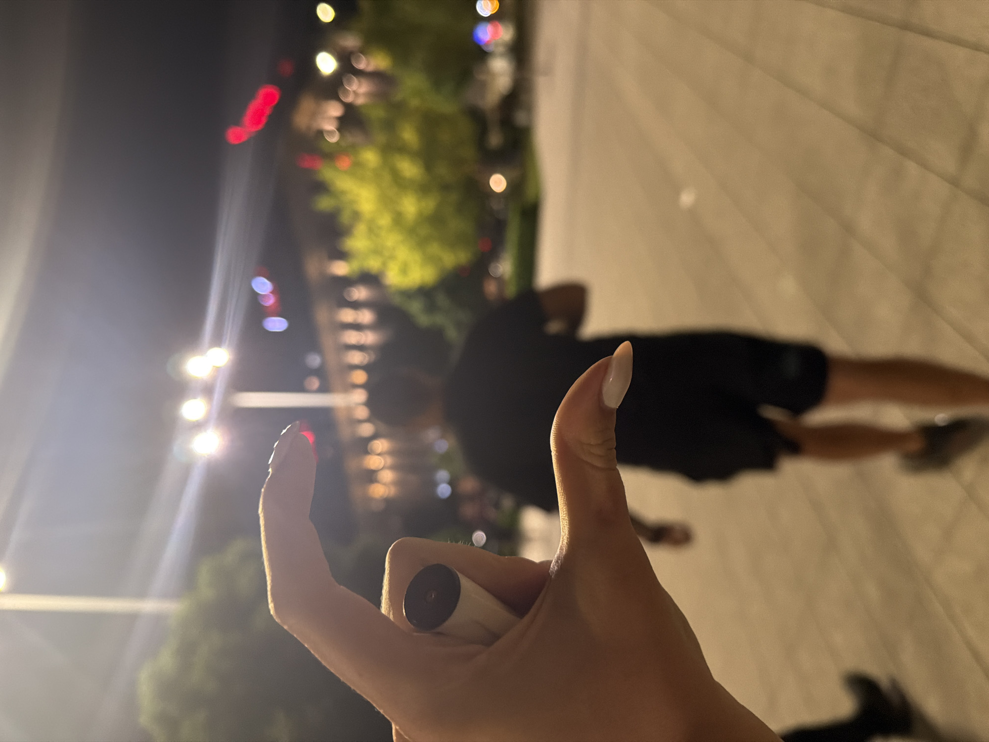 The image, rotated counter-clockwise, captures a night scene with a primary focus on a person's right hand in the foreground, giving a "thumbs up" gesture. The hand features long, light-colored manicured nails. A small, cylindrical, cream-colored object with a dark top, possibly an electronic vape pen, is held between the thumb and forefinger.
In the blurred mid-ground, a person, indistinct due to motion or depth of field, is seen walking away from the camera. This individual appears to be wearing dark clothing, including a t-shirt and shorts. The background is characterized by numerous out-of-focus bokeh lights in various colors (white, red, yellow, blue), suggesting an urban or semi-urban outdoor environment at night. Some blurred green foliage is also visible, hinting at trees or bushes. The ground underfoot appears to be a light-colored paved surface, consistent with a street, promenade, or plaza. The prominent artificial illumination confirms the nighttime setting. The "thumbs up" gesture typically conveys a positive sentiment such as approval or satisfaction. No visible text is present.