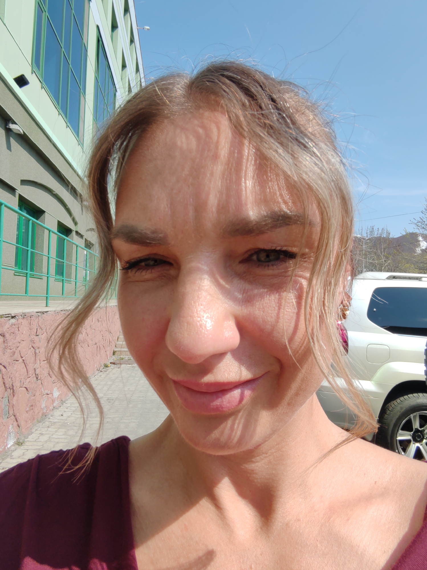 A close-up selfie of a blonde woman with light skin, potentially in her late 30s or early 40s, is taken outdoors on a sunny day. She is slightly smiling and looking at the camera. She appears to be wearing a dark burgundy or dark red top. Her hair is styled with loose waves framing her face.
Behind her is a light green building with rectangular windows with dark blue or green glass. There is a green metal railing or fence running along the side of the building, which is on a raised platform with a stone textured wall below. To the right, a white SUV is partially visible. The sky is clear and blue.
Based on the architectural style of the building and the general landscape, the location is possibly Petropavlovsk-Kamchatsky, Russia.
