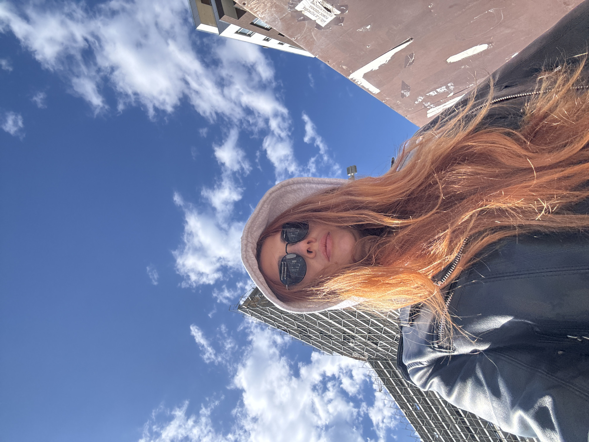 A low-angle perspective captures a woman with red hair wearing sunglasses and a hoodie. She is outside, likely in an urban setting. The background includes a bright blue sky with scattered white clouds, alongside parts of buildings and a steel structure. The location might be Budva, Montenegro.