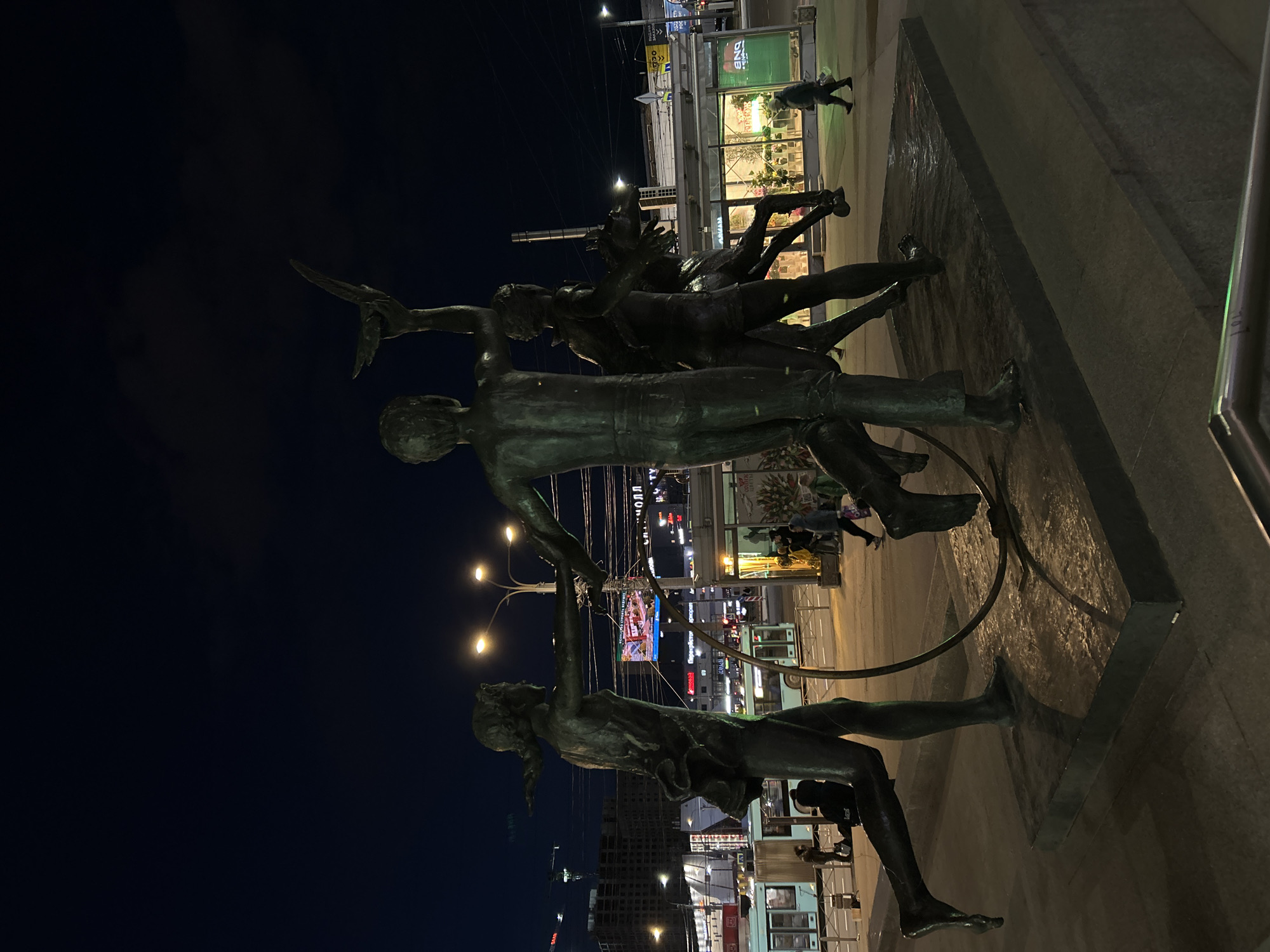 The image captures a dynamic bronze sculpture at night in an urban setting, likely Saint Petersburg, Russia. The sculpture features a group of several muscular human figures, nude or minimally clothed, depicted in active, interconnected poses, suggesting motion, dance, or a collective endeavor. One prominent figure stands upright, while another figure, positioned lower and to the left, appears to be holding onto a circular hoop or ring. A third figure, higher and to the right, extends an arm holding an object resembling a bird or a dart. The dark bronze material of the statues contrasts sharply with the brightly lit city background.
The setting is a lively city street or square illuminated by numerous artificial lights. In the background, buildings glow with various signs and windows, and streetlights cast warm light onto the scene. Distant traffic and people walking on sidewalks are visible. A large digital billboard in the distance displays advertisements with Cyrillic text, including "РЕДМОНД" (Redmond) and "СПОРТМАСТЕР" (Sportmaster). Other discernible signs include a green sign with "DND" or "OND" and a yellow "ВЫХОД" (Exit) sign. The sculpture rests on a dark, reflective base, possibly a water feature, which subtly mirrors the surrounding lights. The overall atmosphere is energetic and urban, with the dramatic night lighting emphasizing the sculptural forms.