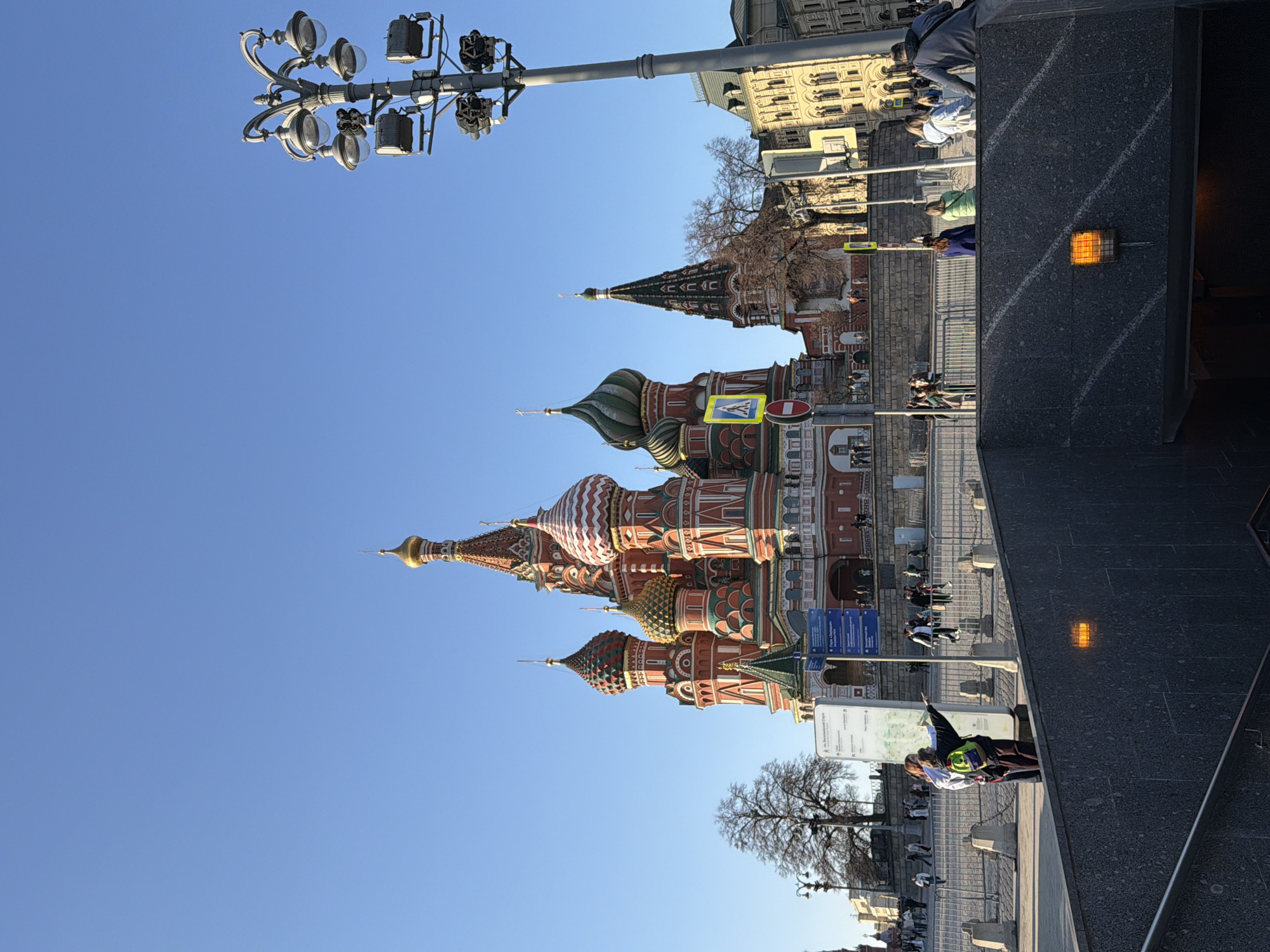The image features St. Basil's Cathedral in Moscow, Russia, under a clear blue sky. The cathedral's colorful domes are prominently displayed. A street lamp with multiple lights stands to the left of the cathedral. Pedestrians are visible along the street. In the foreground, part of a gray structure, possibly a building entrance, is visible, with an orange light fixture on its wall.