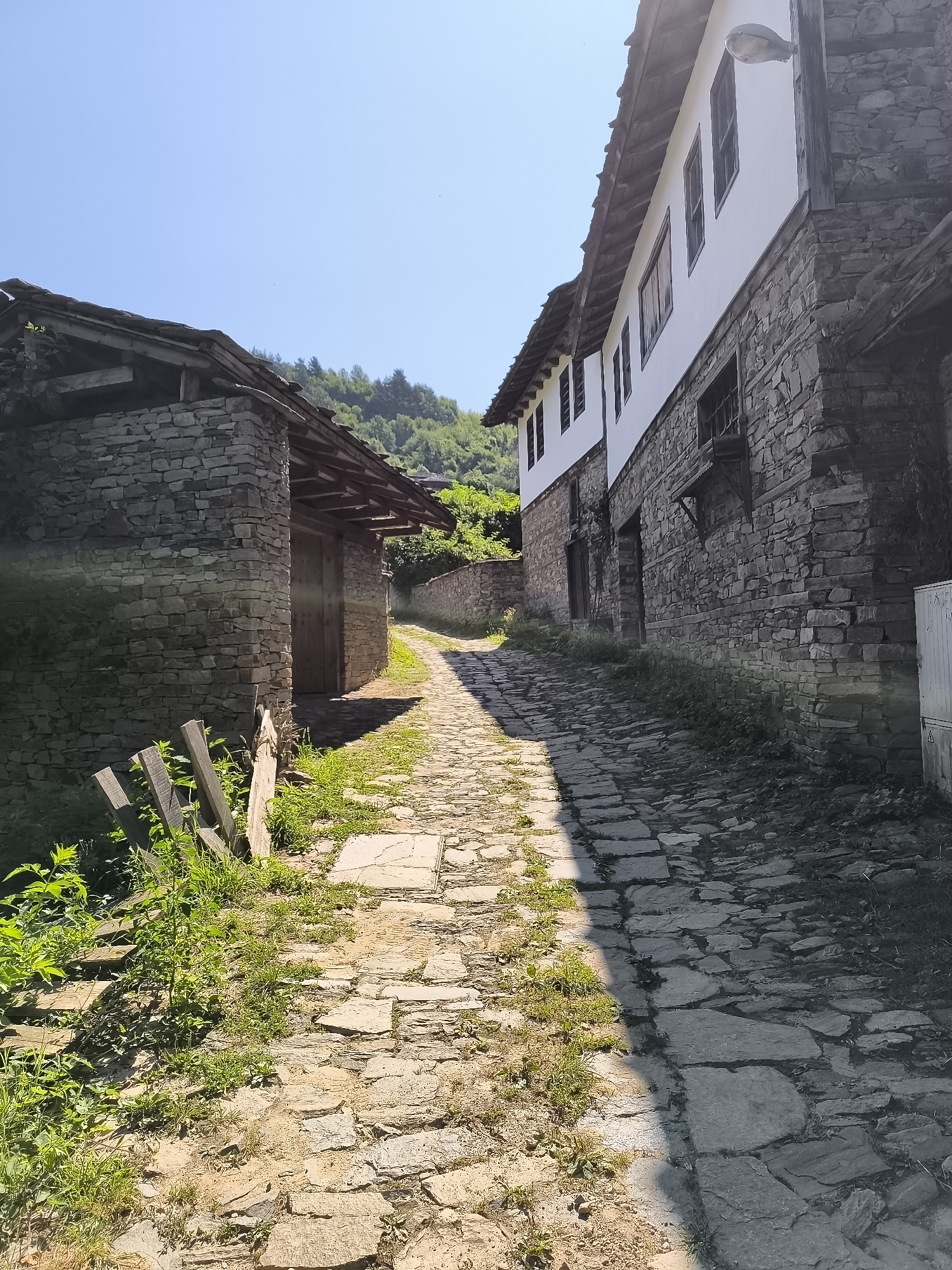 The image depicts a narrow, stone-paved street in the historic village of Kovachevitsa, Bulgaria. On the right, a traditional Bulgarian house features a white upper story with dark-framed windows, contrasting with a lower story constructed of rough-hewn stone. The roofline of this house is visible, with a dark, tiled or shingled covering. To the left, a smaller, single-story stone building with a wooden roof is situated. A wooden gate or fence section is visible in the foreground to the left of the street. The street itself is uneven, paved with irregularly shaped stones, with patches of grass and dirt growing between them. Sunlight casts a sharp shadow across the right side of the street, indicating a sunny day. The background shows a lush, green hillside with trees, and a clear blue sky above. The overall impression is of a quiet, old-world village setting.