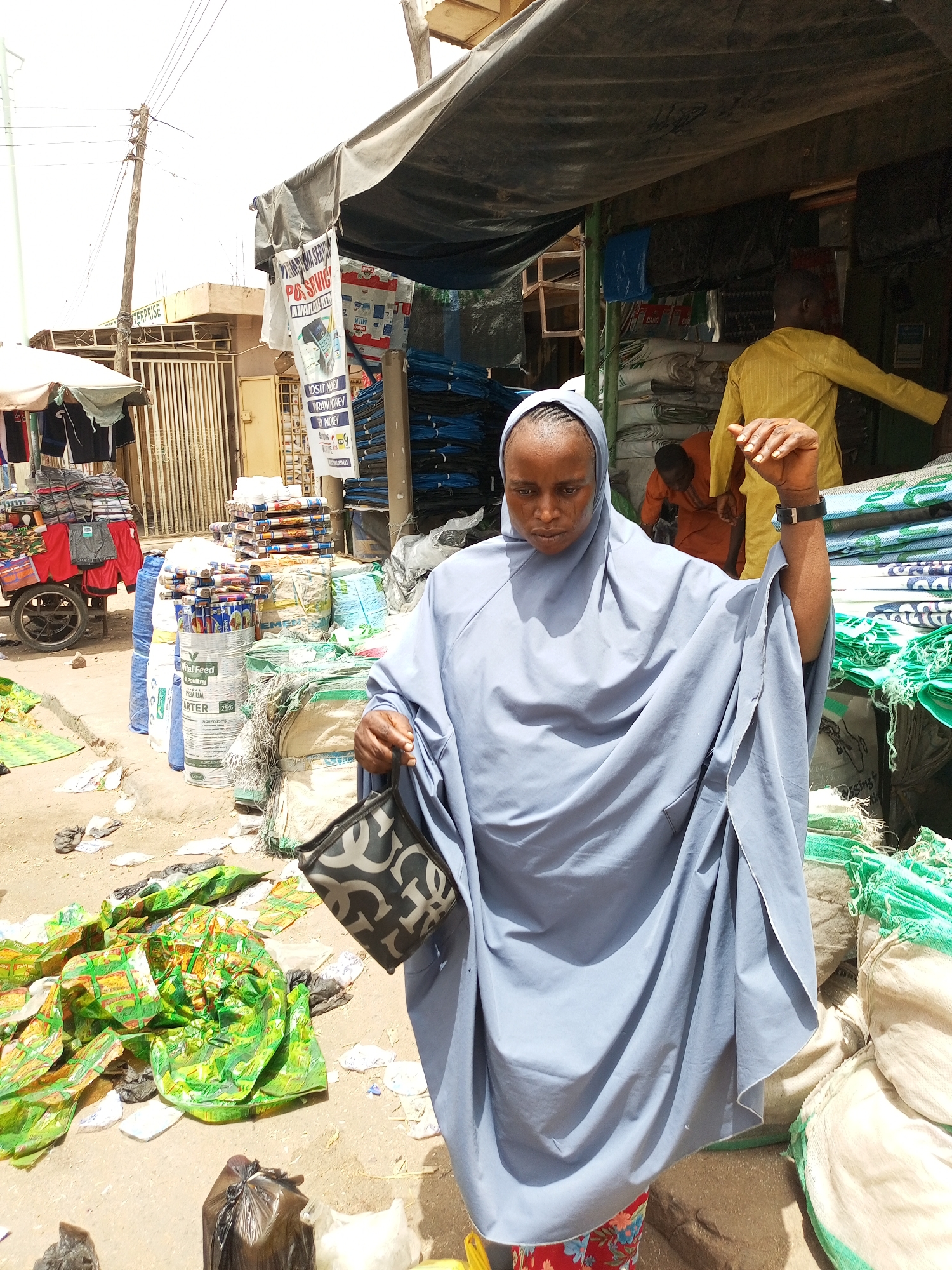 A medium shot depicts a woman in Arawa, Nigeria. She wears a blue headscarf and a matching robe-like garment, holding a black and gray handbag. She stands amidst a bustling outdoor market. Behind her are sacks of goods, and in the background, two men are partially visible under a makeshift shelter or stall. The market environment includes various items for sale and litter scattered on the ground. A sign on a nearby building is partially visible, advertising services and listing products. An electric pole is also visible.