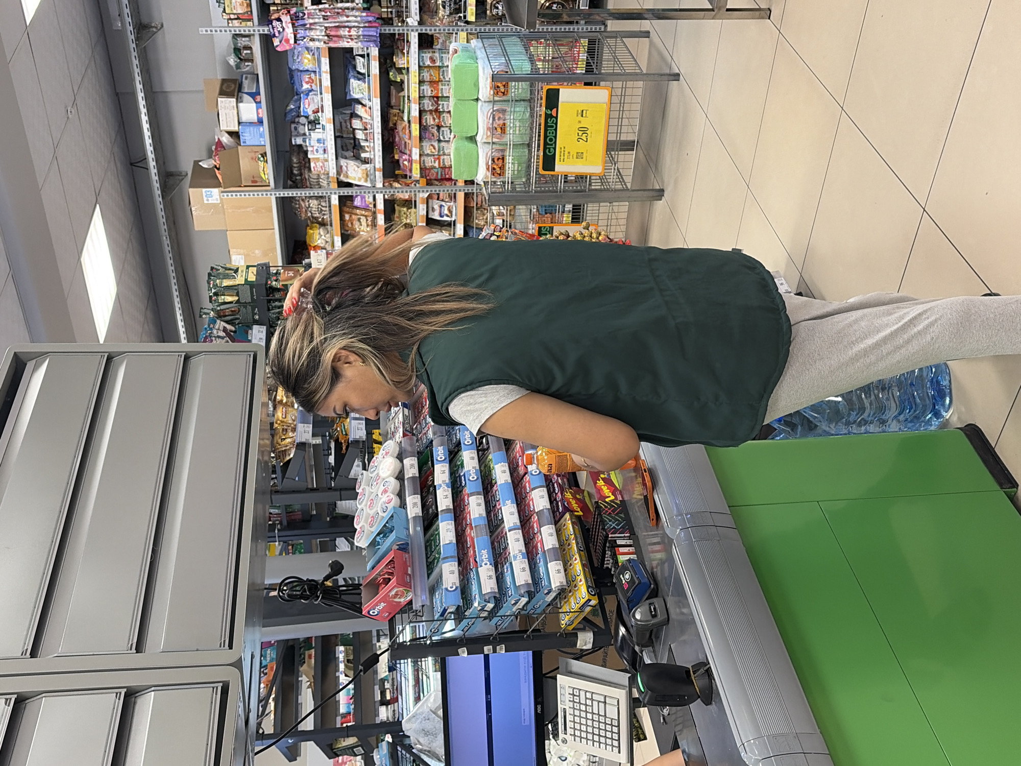 An adult female with long, light brown hair is standing at a grocery store checkout counter, facing away from the camera. She is wearing a dark green vest over a white t-shirt and light gray sweatpants. Her right hand is in her hair, and her left hand is near her side. A large blue water jug is visible on the floor to her right. The checkout counter is green and gray, with a computer monitor, calculator, barcode scanner, and various small items on it. Behind her, shelves are stocked with packaged goods, including chewing gum and cleaning supplies. A shopping cart with a yellow sign displaying "GLOBUS" and the price "250" is to her right, filled with green-wrapped items. The floor is tiled with a beige and white checkerboard pattern. The lighting suggests an indoor environment, likely a supermarket.