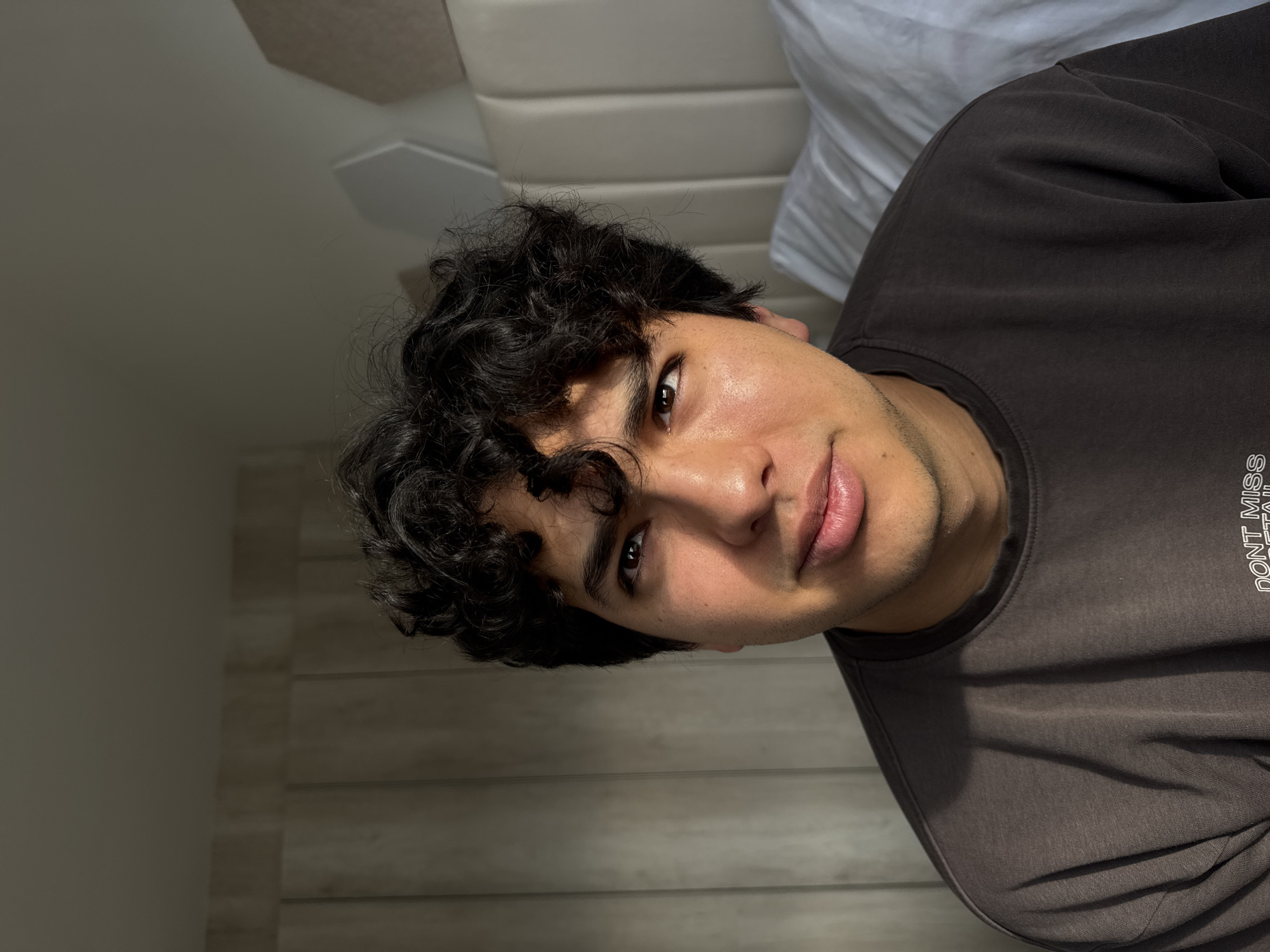 A person with dark curly hair and light-brown skin is looking at the camera with a slight smile. The person is wearing a dark t-shirt with the words "DONT MISS." Behind the person, there is a white headboard and part of a white sheet. The background wall appears to be lined with narrow, horizontal panels. The photograph is slightly rotated counter-clockwise. The location is not discernible from this image.