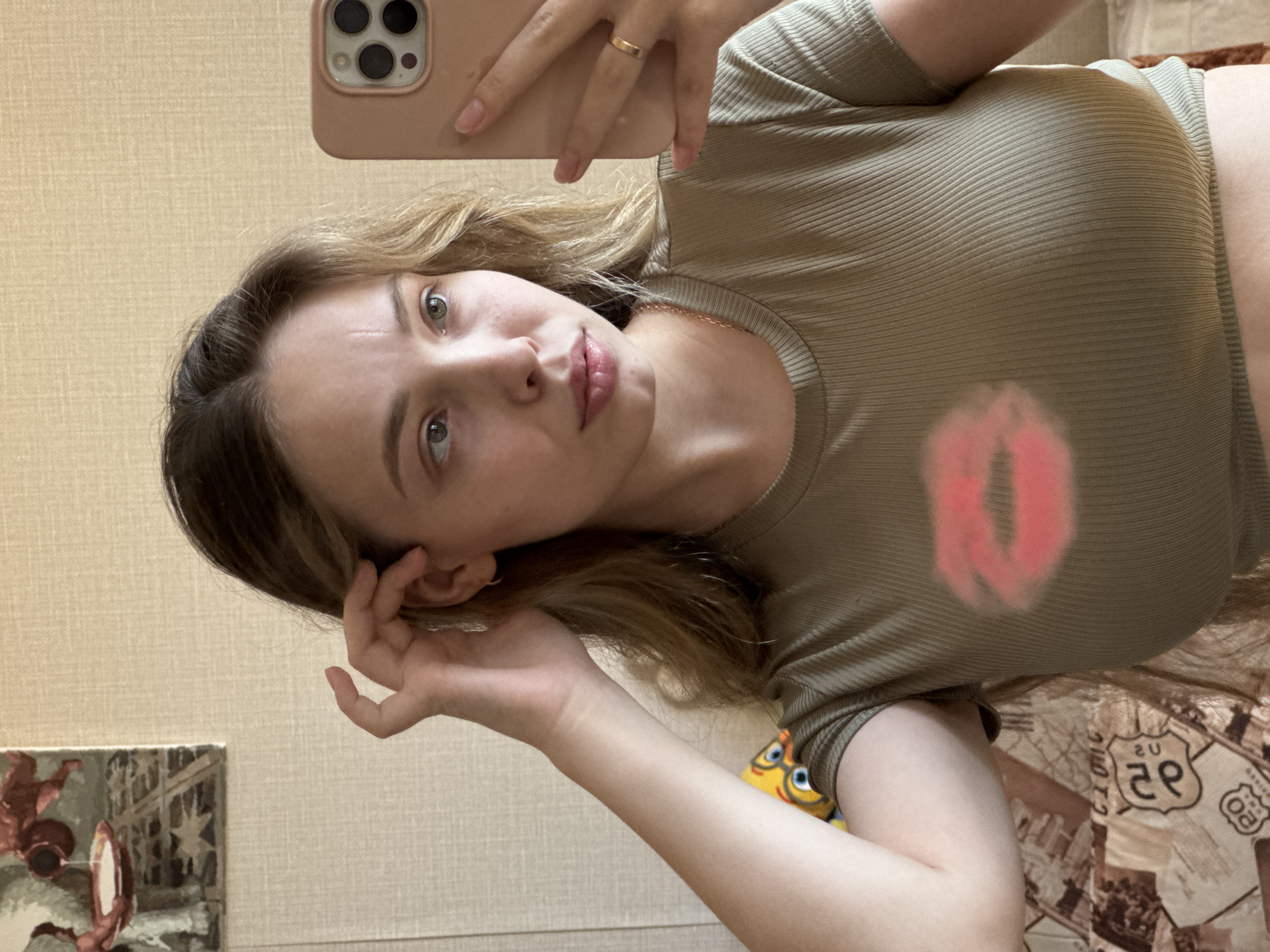 A young woman takes a selfie with a phone, holding it in her right hand. She has blonde hair, fair skin, and appears to be wearing a gold ring on her ring finger. She is wearing a ribbed, olive-green cropped t-shirt with a pink lipstick kiss mark on the front. Her left hand is near her ear. The background shows a cream-colored wall, a picture frame with a landscape painting, and a bed covered in a printed sheet with images related to Route 66. The location is possibly an interior in the city of Chaikovsky, Russia.