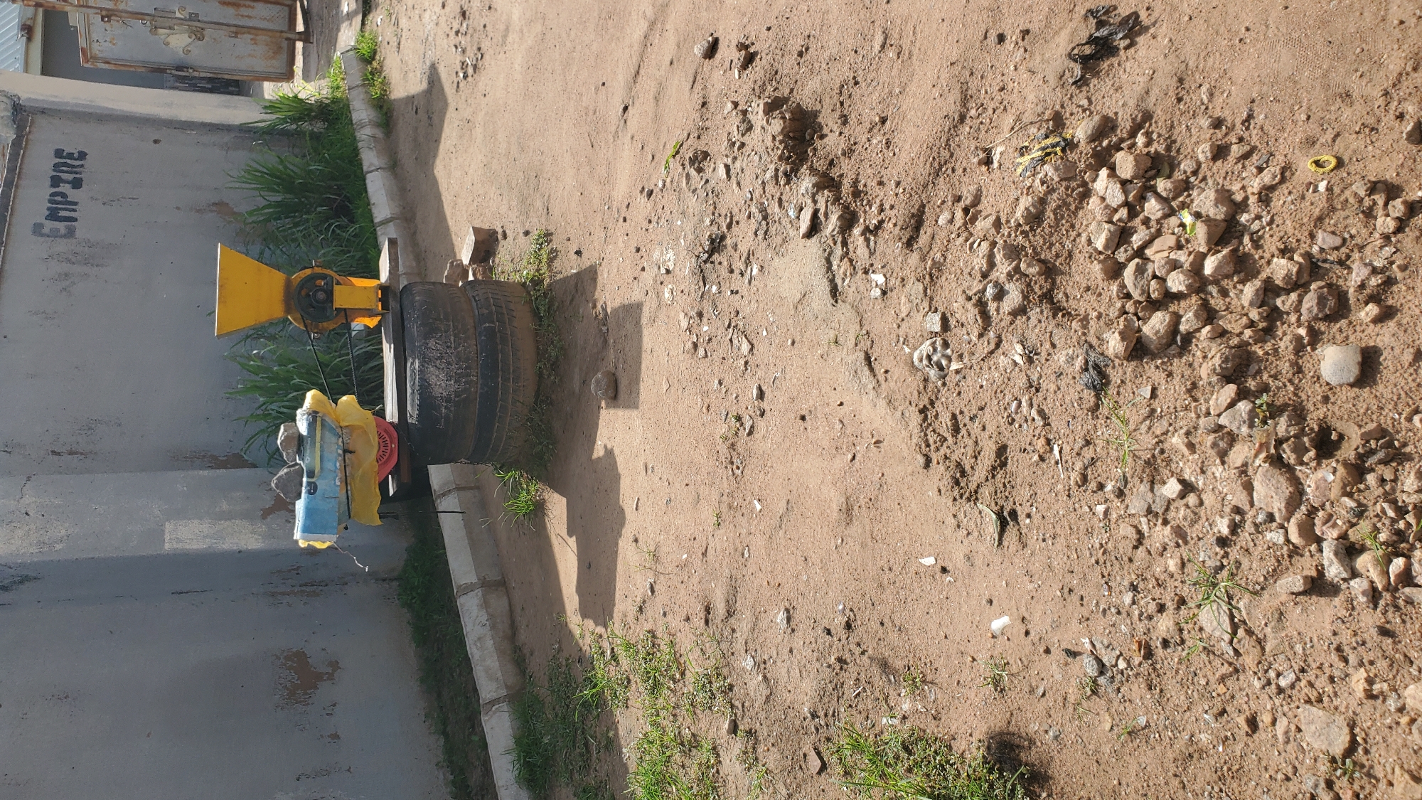 The image depicts an outdoor scene in Jalingo, Nigeria, with a focus on a makeshift contraption. The ground is a mixture of dirt and gravel, with scattered rocks and sparse patches of grass. In the background, a weathered concrete wall is visible, with the word "EMPIRE" spray-painted on it. Adjacent to the wall is a doorway with a rusty metal gate.
The central object is a grain mill or grinder, characterized by a yellow hopper on top. This mill is mounted on two stacked tires, which serve as a base. Beneath the mill and tires, a wooden plank is used for support, placed on top of some bricks. Resting on top of the mill's mechanism is a blue object wrapped in yellow plastic, with two rocks placed on it. A belt connects the mill's pulley system to another component, likely part of a motor or power source, though the source itself is not clearly visible.
The overall environment suggests a semi-rural or industrial area, possibly a workshop or storage space, with evidence of makeshift repairs and repurposed materials. The lighting indicates it is daytime, with shadows cast by the objects.