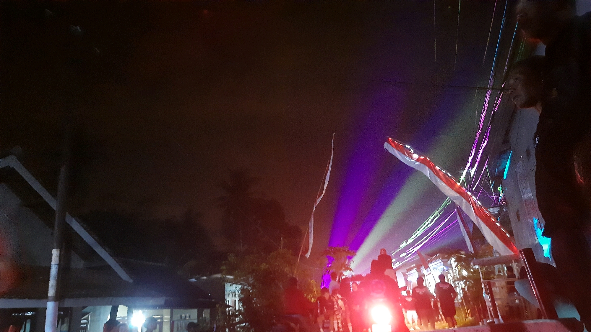 The image captures an outdoor nighttime scene, possibly in the Kebondalem area of Indonesia, characterized by artificial lighting and a gathering of people. In the foreground, the bright headlights of a motorcycle illuminate a group of individuals walking or standing. Several people are visible, some appearing to be moving towards the camera, while others are further back.
The background is dominated by a dark sky with diffused light, suggesting a humid or cloudy atmosphere. Several structures are present, including what appears to be the roof of a building with angled supports. String lights are strung across the area, some emitting a vibrant purple and green hue, contributing to the festive or event-like atmosphere. A long, thin object, possibly a flag or banner, hangs vertically in the middle ground, its colors not clearly discernible due to the lighting conditions. To the right, a portion of a red and white flag, likely the Indonesian flag, is prominently displayed, draped across a structure and illuminated by spotlights. A person is visible in the upper right corner, looking towards the event. The overall impression is one of a lively nighttime event, possibly a celebration or festival, with a strong visual emphasis on colored lights and national symbols.