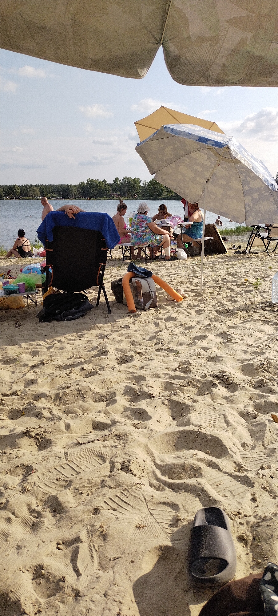 This image depicts a sunny day at a beach by a body of water, likely Lake Bezliudivske in Bezliudivka, Ukraine. In the foreground, on the sandy beach, there are several items including a dark folding chair with a blue towel draped over it, a backpack with orange pool noodles, and a single dark slide sandal. Further back, a group of people are gathered around a table under a large patterned umbrella. Some individuals are seated, eating or conversing, while others appear to be standing or in the water. The water's edge is visible, with trees forming a treeline in the background. The overall scene suggests a recreational outing.