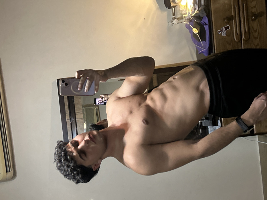 A shirtless man with curly dark hair stands in front of a mirror, holding a phone to take a picture of his reflection. He is wearing black shorts or pants. The background includes a beige wall, an air conditioning unit, and a wooden cabinet with a sticker on it. The lighting is somewhat dim, creating shadows on the man's body. A power strip and additional items are visible on the cabinet.