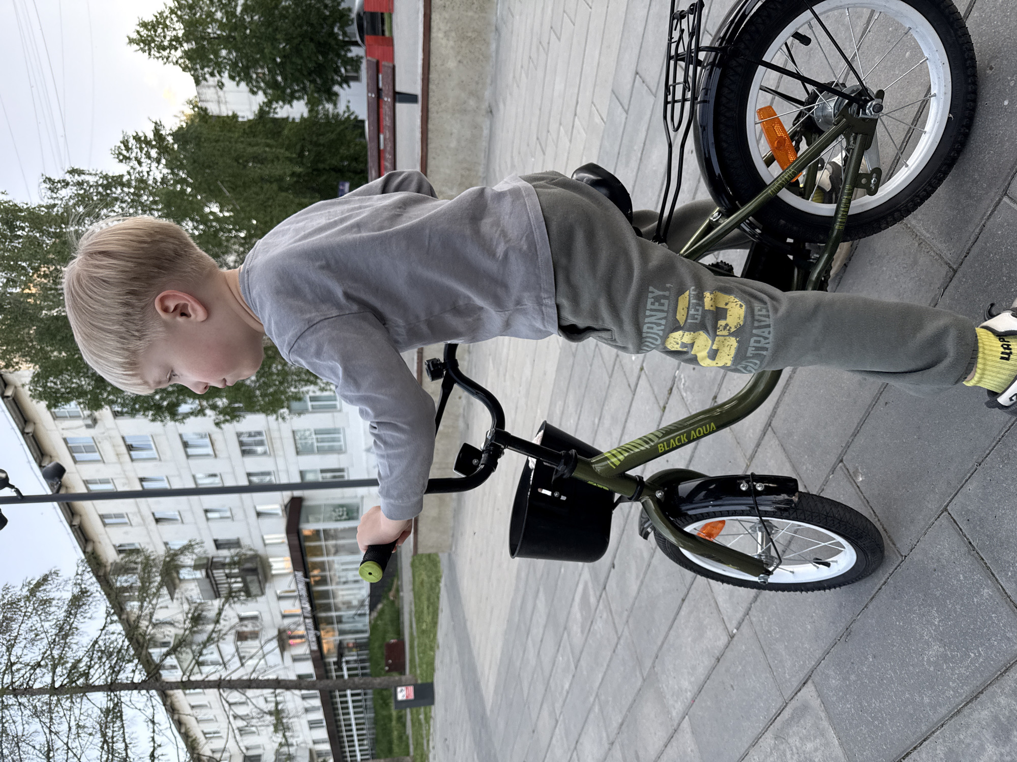 A young boy is leaning over the handlebars of a green "Black Aqua" bicycle on a paved surface. The boy is wearing a gray long-sleeved shirt and gray sweatpants with the words "Journey" and "Let's Travel" printed on the pant leg. He has blonde hair. The bicycle has a black front basket, white rims on the tires, and a rear rack. A multi-story building is visible in the background, and the location appears to be an urban setting. While the question asks for the location to be the city of Chaikovsky, Russia, this cannot be conclusively confirmed from the image alone.