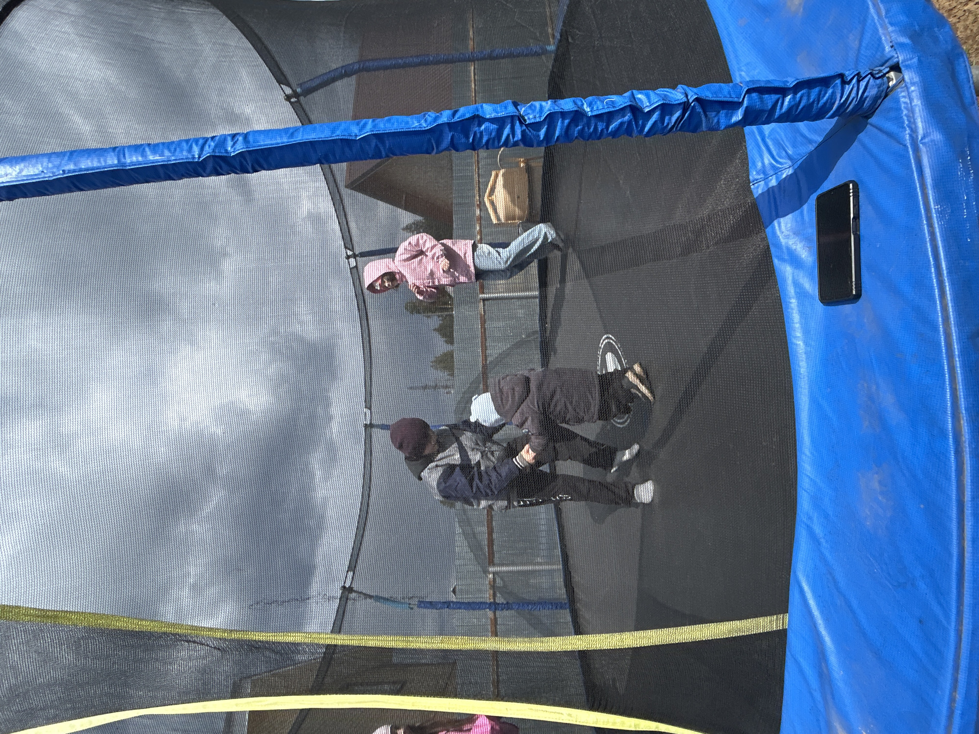 Two children are inside a trampoline with a safety net enclosure. One child, wearing a pink hooded jacket and blue jeans, is standing near a tan-colored bag hanging from the net. The second child, wearing a dark jacket, pants, a hat, and light-colored sneakers, is positioned closer to the trampoline's center. A mobile phone is seen attached to the blue safety pad surrounding the trampoline's edge. The trampoline is outdoors, and a cloudy sky is visible through the net. The exact location within Chaikovsky, Russia cannot be determined from this image.